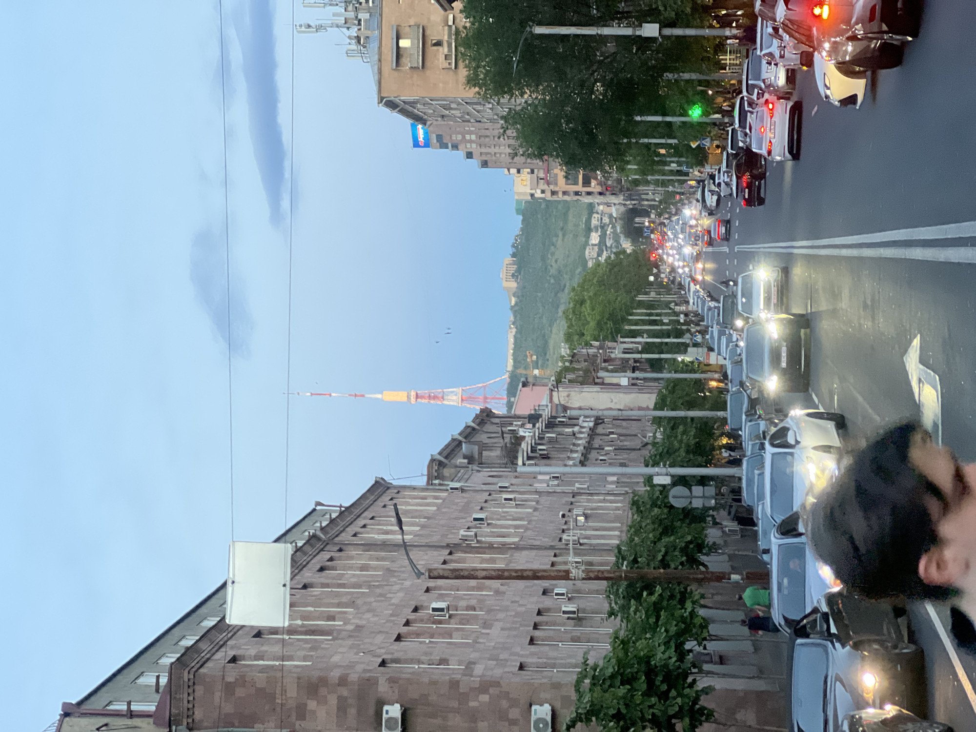 The image depicts a street scene in Yerevan, Armenia, with the Yerevan TV Tower visible in the distance. The photo is taken from street level and shows a long avenue filled with cars, creating a traffic jam. The buildings lining the street are a mix of older, multi-story stone structures and more modern ones. Many of the older buildings are equipped with numerous air conditioning units. Trees are planted along the sidewalk, partially obscuring the buildings. The sky is overcast with a light blue hue. A person's face and hair are visible in the lower right corner, possibly the photographer.