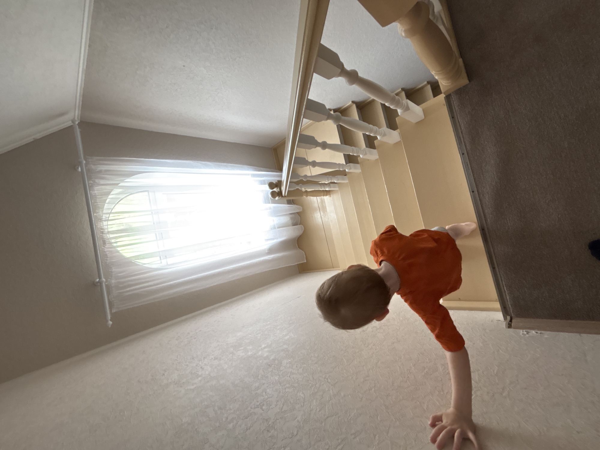 A child in an orange shirt is upside down in a room. The room has a window with white curtains, beige walls, and light-colored flooring. A staircase with white banisters is also visible. The location is Ферма, Russia.