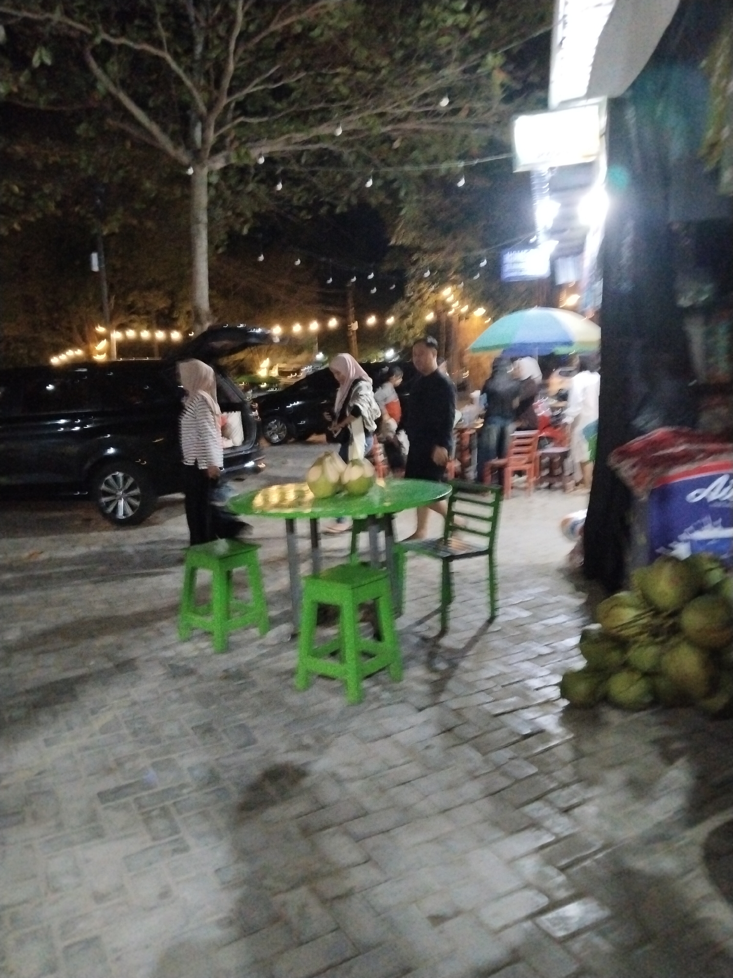This is a nighttime, low-light photograph taken at a street-side food stall in Prigi, Indonesia. The scene is outdoors on a paved surface, likely a sidewalk or parking area, with a tree overhead. In the foreground, a green table with a matching chair and two stools are arranged. On the table are at least two green coconuts. To the right of the table, a pile of green coconuts is visible.
There are several people in the image. A person wearing a light-colored headscarf and a striped shirt is standing near an open black SUV. Another person, also wearing a headscarf, stands to their right, holding something. In the background, further people are visible near a colorful umbrella and some tables and chairs, suggesting a small seating area for customers. The overall lighting is dim, with string lights visible in the background. The presence of coconuts and the setting suggests this is a place where coconuts are being sold or served, likely as a refreshment.