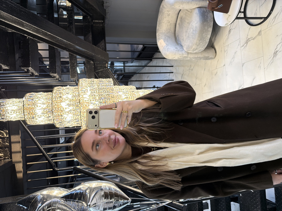 A young adult female is depicted in a mirror selfie taken indoors in Moscow, Russia. She holds a light-colored smartphone with a triple-lens camera in her right hand, positioned in front of her face. The woman has long, light brown hair, a faint smile, and wears a dark brown long coat, a brown turtleneck, a cream-colored scarf, a gold cross pendant, pearl drop earrings, and gold rings. Her fingernails are painted light pink. The background features a tall, multi-tiered crystal chandelier illuminating the space. To the left, several reflective silver balloons are partially visible near a dark metal staircase railing. To the right, a dark wooden or metal staircase ascends. A light grey, textured armchair with a curved design is situated on a light marble floor with grey veining. A small round side table with a white marble top and black metal base holds a dark brown leather handbag with a top handle and gold hardware. The environment suggests an upscale interior setting.