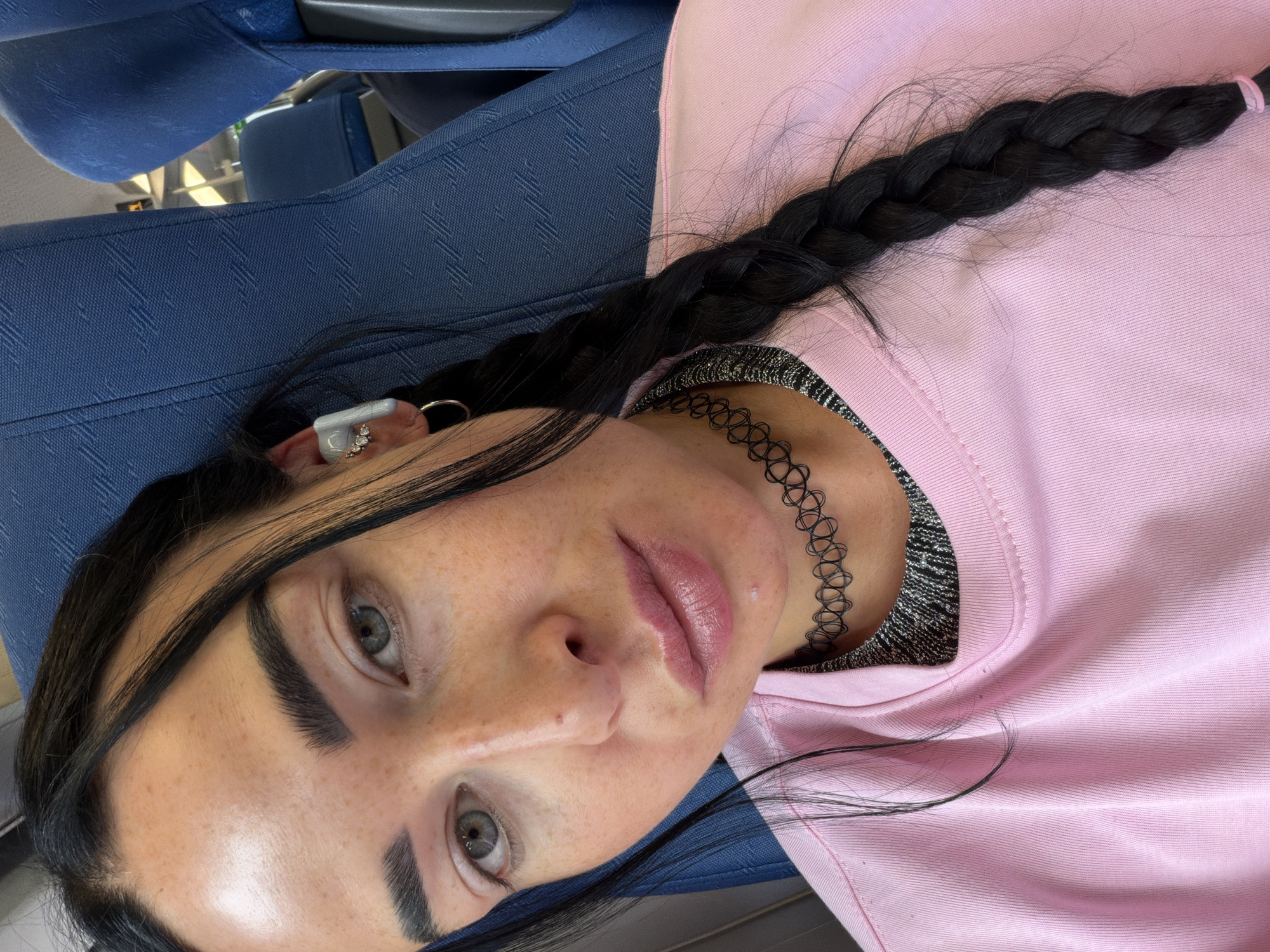 This media file shows a close-up, overhead shot of a young woman lying down on a blue seat, likely on public transportation given the texture and pattern of the seat. She is wearing a pink sweatshirt over a black and silver metallic top and a black choker necklace. Her long, black hair is styled in a braid that rests on her shoulder and the sweatshirt. She has striking light green eyes, dark eyebrows, and a scattering of freckles across her nose and cheeks. Her lips are full and appear to have a pinkish tint. She is wearing an ear cuff with small stones in her left ear. The lighting suggests it is daytime. The scene appears to be indoors, possibly in transit in or around Ушаки, Russia. There is no visible text in the image.