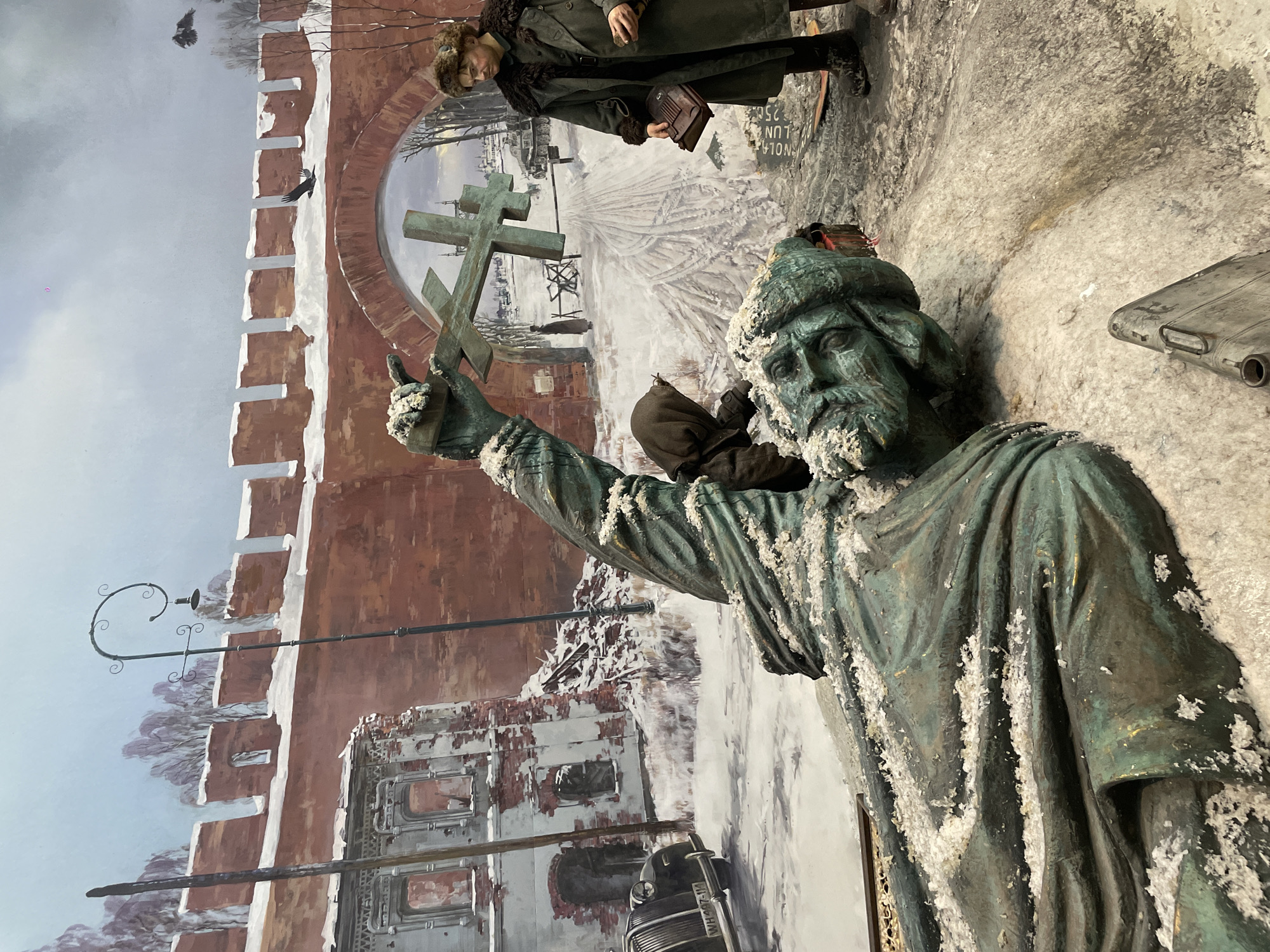 The image depicts a statue of a man holding a cross, covered in what appears to be snow. The statue stands in front of a mural that includes a depiction of the Moscow Kremlin wall and snowy street scenes. A person wearing a winter coat and hat is also visible near the upper part of the mural. A car with the license plate “WH-47-11” appears at the bottom of the mural.