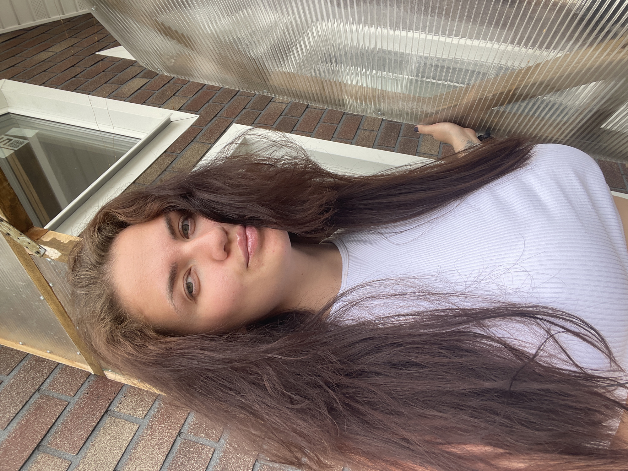 A young woman with long, brown hair is lying on what appears to be a brick surface with a white ribbed shirt on. A window is visible behind her and a clear corrugated plastic panel behind that. The location is potentially Мосметаллоконструкция Онт, Russia.