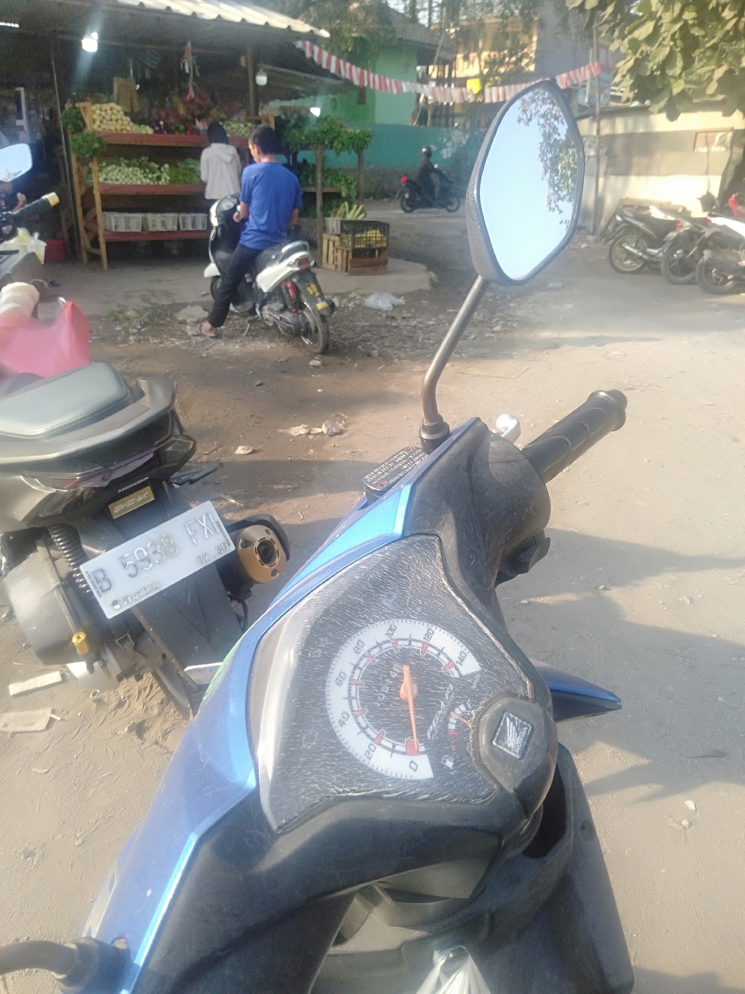 In Sukadami, Indonesia, a street vendor stall is visible in the background, displaying fruits and vegetables. A young man in a blue t-shirt is seated on a white scooter in front of the stall. Another person, wearing a white hoodie, stands near the stall. Several other scooters are parked nearby. The foreground is dominated by the front of a blue scooter, its dashboard and handlebars clearly visible. The image captures a sunny day with shadows cast on the sandy ground.