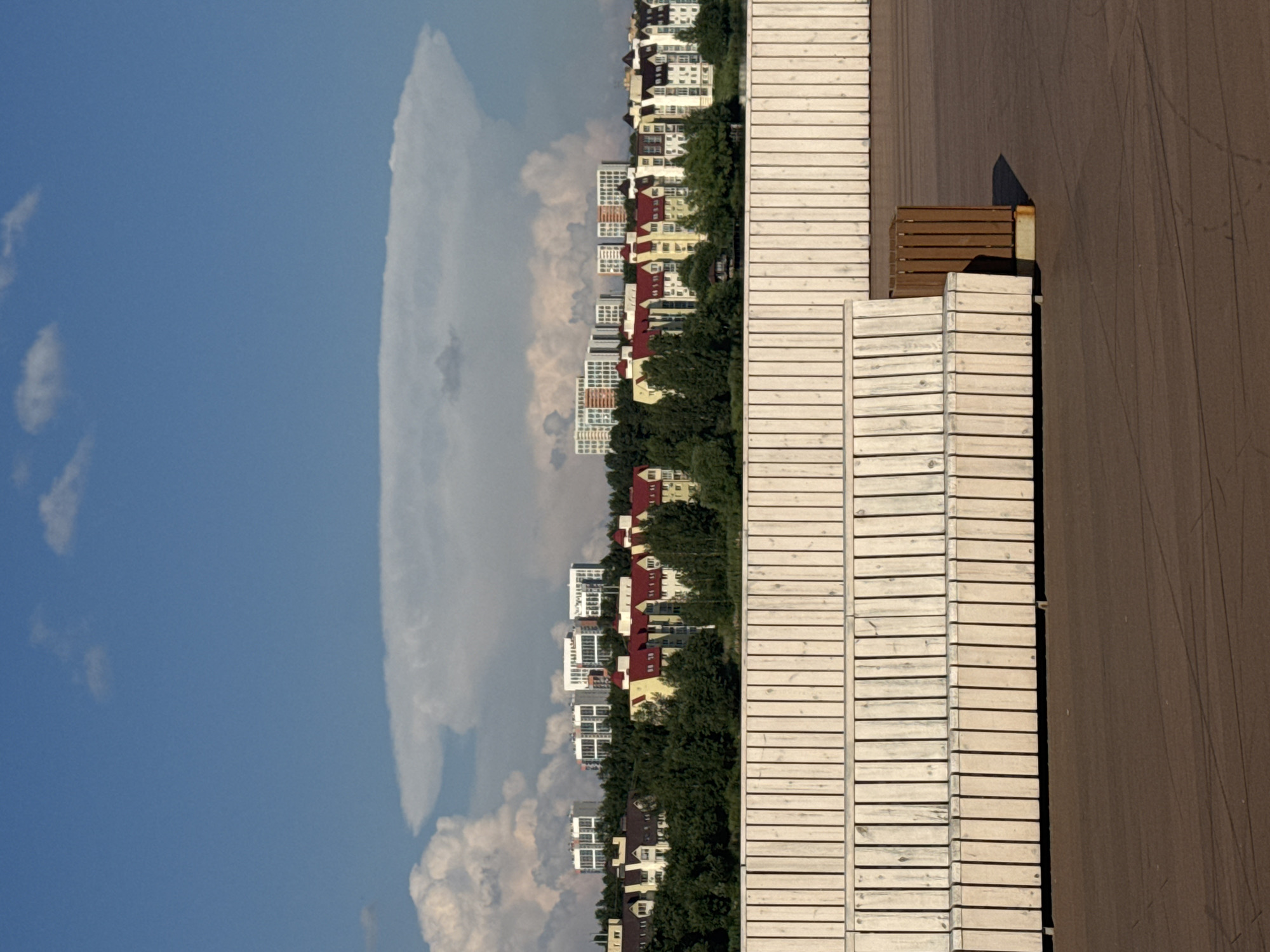 The image depicts a waterfront promenade in Yekaterinburg, Russia. In the foreground, a modern wooden bench with a trash receptacle is situated on a dark brown, textured surface, possibly asphalt or concrete. Behind the bench is a long, light-colored wooden fence, extending horizontally across the frame.
In the middle ground, a line of trees separates the promenade from a cityscape of residential buildings. The buildings vary in architectural style and color, with some featuring red roofs and others a more modern white facade. The buildings appear to be apartment complexes.
The background is dominated by a vast, blue sky with scattered white clouds. A prominent, large anvil-shaped cloud formation is visible, characteristic of a cumulonimbus cloud. The overall impression is a clear, sunny day with a dramatic cloud formation. The scene is tranquil and urban, with no people visible.