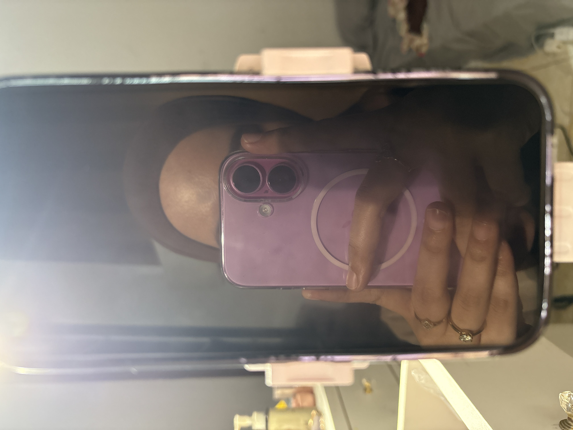 A selfie taken with a purple iPhone is reflected in a makeup mirror, presumably by a person whose face and torso are partially visible. The hand holding the phone has two rings on two different fingers. Below the mirror, portions of vanity items are partially visible. The setting is unknown and there is no indication of the city of Gadingrejo, Indonesia.