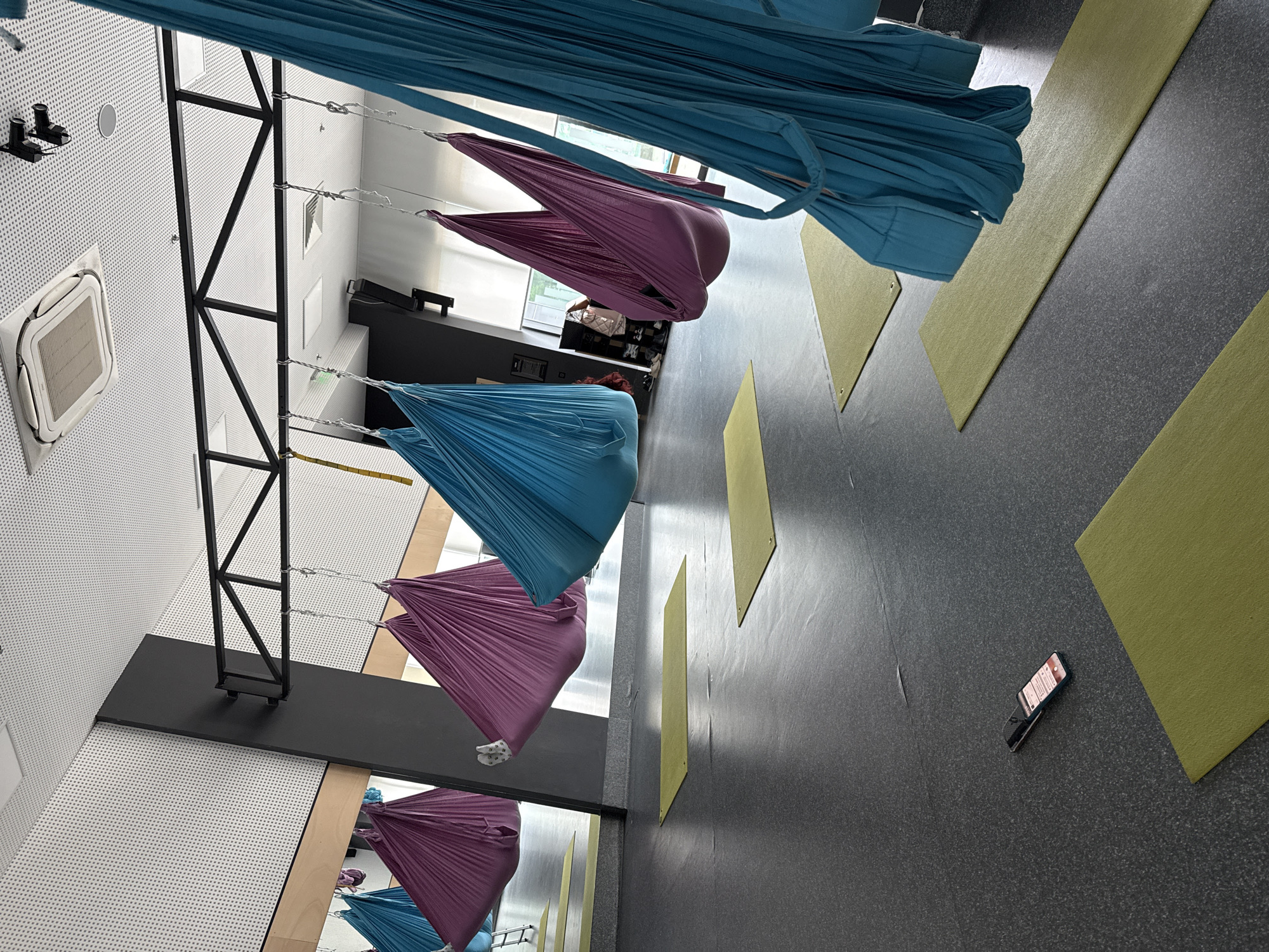 The image captures an interior space, likely a yoga or aerial fitness studio in Unknown, Unknown. Several aerial yoga hammocks in alternating colors of blue and purple hang from a black metal frame suspended from the ceiling. Exercise mats are placed on the floor. A cell phone is lying on the floor between two mats. A mirror is located along a wall.