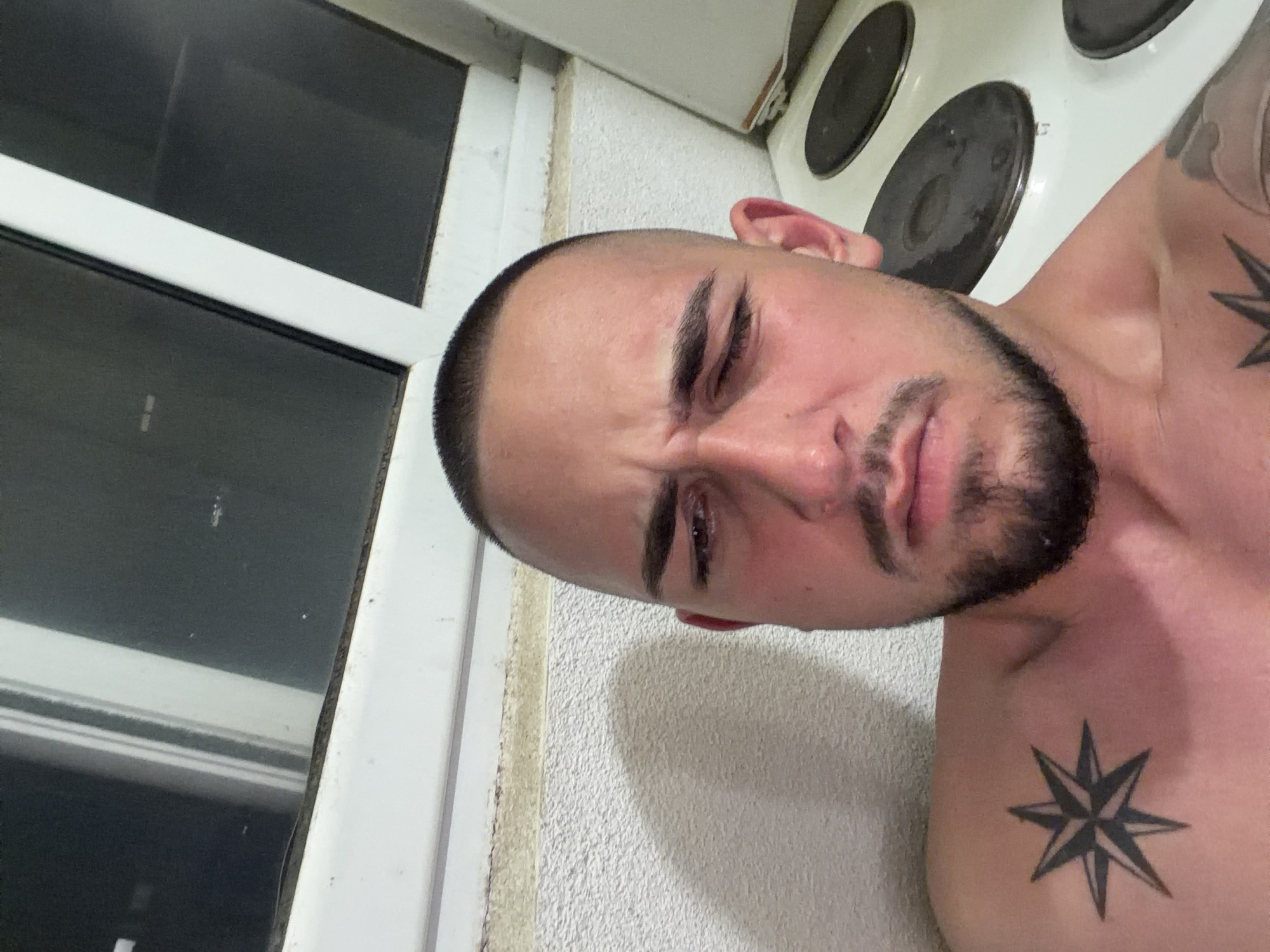 This image depicts a male individual taking a selfie indoors, in Sofia, Bulgaria. The subject is a light-skinned adult male with a very short haircut, dark eyebrows, and a short beard. He is shirtless, revealing an eight-pointed star tattoo on his left clavicle/shoulder and a partial tattoo on his right upper arm. His facial expression is a grimace, with his right eye squinted more acutely than his left, and his mouth slightly contorted. The background features a textured white wall, above which a white window frame with dark panes is visible. Below the window frame, a horizontal band of discolored material, consistent with mold or dirt, runs along the wall. To the right of the subject, a white electric stovetop with two dark circular burners is partially visible. The overall scene suggests a domestic setting.