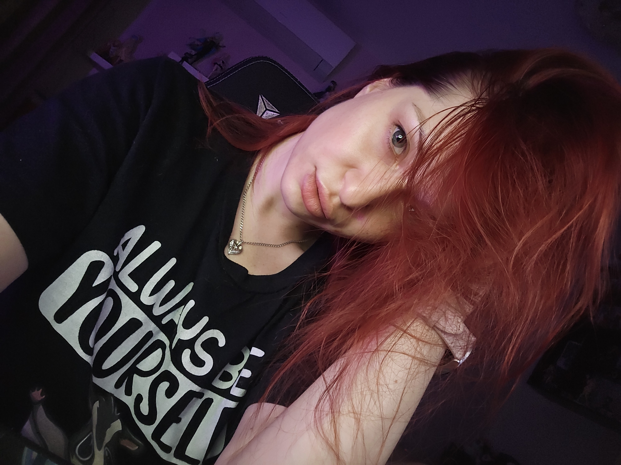 The image displays a Caucasian female with fair skin, light blue-grey eyes, and voluminous reddish-orange hair, situated indoors in Burgas, Bulgaria. Her hair is styled with significant volume, with strands falling across the right side of her face, partially obscuring her right eye. She is wearing a black crew-neck t-shirt featuring the white text "ALWAYS BE YOURSELF" printed across the chest. A thin silver chain necklace with a small, ornate heart-shaped pendant is visible around her neck. Her right arm is raised, with the hand positioned behind her head, mostly concealed by her hair. She is seated in a dark-colored gaming chair, identifiable by a prominent white or silver logo resembling a stylized pyramid or 'S' on the headrest. The environment is dimly lit with a dominant purple ambient light source originating from the left side of the frame, illuminating the wall and part of the room. The background beyond the subject and chair is indistinct, showing dark areas consistent with an interior setting. The perspective suggests a self-portrait taken from a slightly low angle.