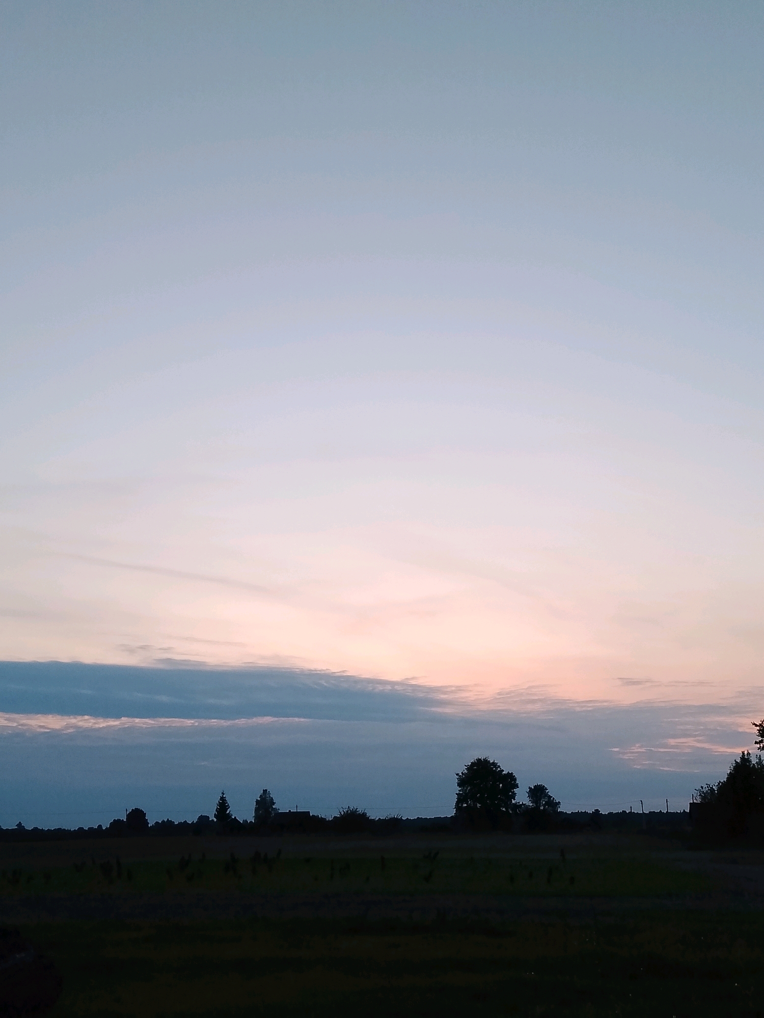 The image depicts a rural landscape at dusk or dawn, with the sky dominating the upper portion of the frame. The sky exhibits a gradient of colors, transitioning from a pale blue at the top to soft pinks and oranges closer to the horizon, indicating twilight. Several wispy clouds are present, some appearing darker and more defined near the horizon.
The lower third of the image captures a silhouetted treeline against the colorful sky. The treeline consists of various trees, some appearing taller and denser than others, suggesting a mix of deciduous and coniferous species. Interspersed within the treeline are faint outlines of structures, possibly houses or farm buildings, and utility poles. The land in the foreground is dark and indistinct, likely a field or meadow, with the detail obscured by low light conditions. The overall impression is one of tranquility and the vastness of the natural environment under a changing sky. The image does not contain any visible people or animals. The location is specified as Ленінскі сельскі Савет, Belarus.