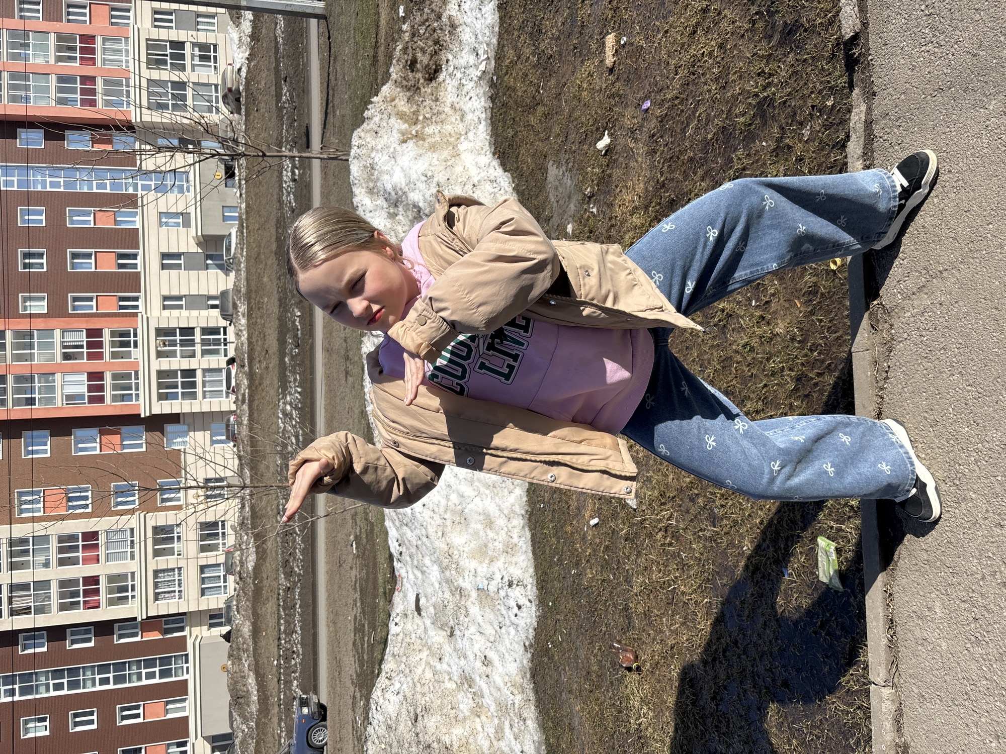 A blonde-haired young girl, dressed in a pink sweatshirt under a beige jacket and patterned blue jeans, stands outside in the daylight. She is on a paved surface, likely a sidewalk. Behind her is a partially melted snow bank and a dirt area. A multi-story apartment building is in the background. A bare tree stands nearby. A small blue car is visible in the background. The location is likely Pervomaysky, Russia, based on the user's prompt. The girl is striking a pose.