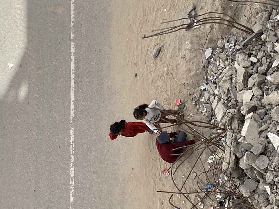An overhead image in Khan Yunis, Palestinian Territory, depicts three individuals on a sandy ground adjacent to a grey asphalt road with a faded white dashed line. In the foreground, a large pile of concrete rubble mixed with rusted, bent rebar is visible. One person, wearing a dark red long-sleeved top, blue pants, and a light blue head covering, is kneeling on the sand, facing downwards and interacting with the ground near the rubble. A young girl in a white long-sleeved shirt with a black "CHANEL" logo, brown patterned trousers, and sandals stands beside the kneeling individual, placing her right hand on their head. A second young child in a red long-sleeved top stands slightly behind the girl, looking towards the left with one hand near their forehead. Scattered small items of debris, including plastic pieces, are present on the sand and within the rubble.
