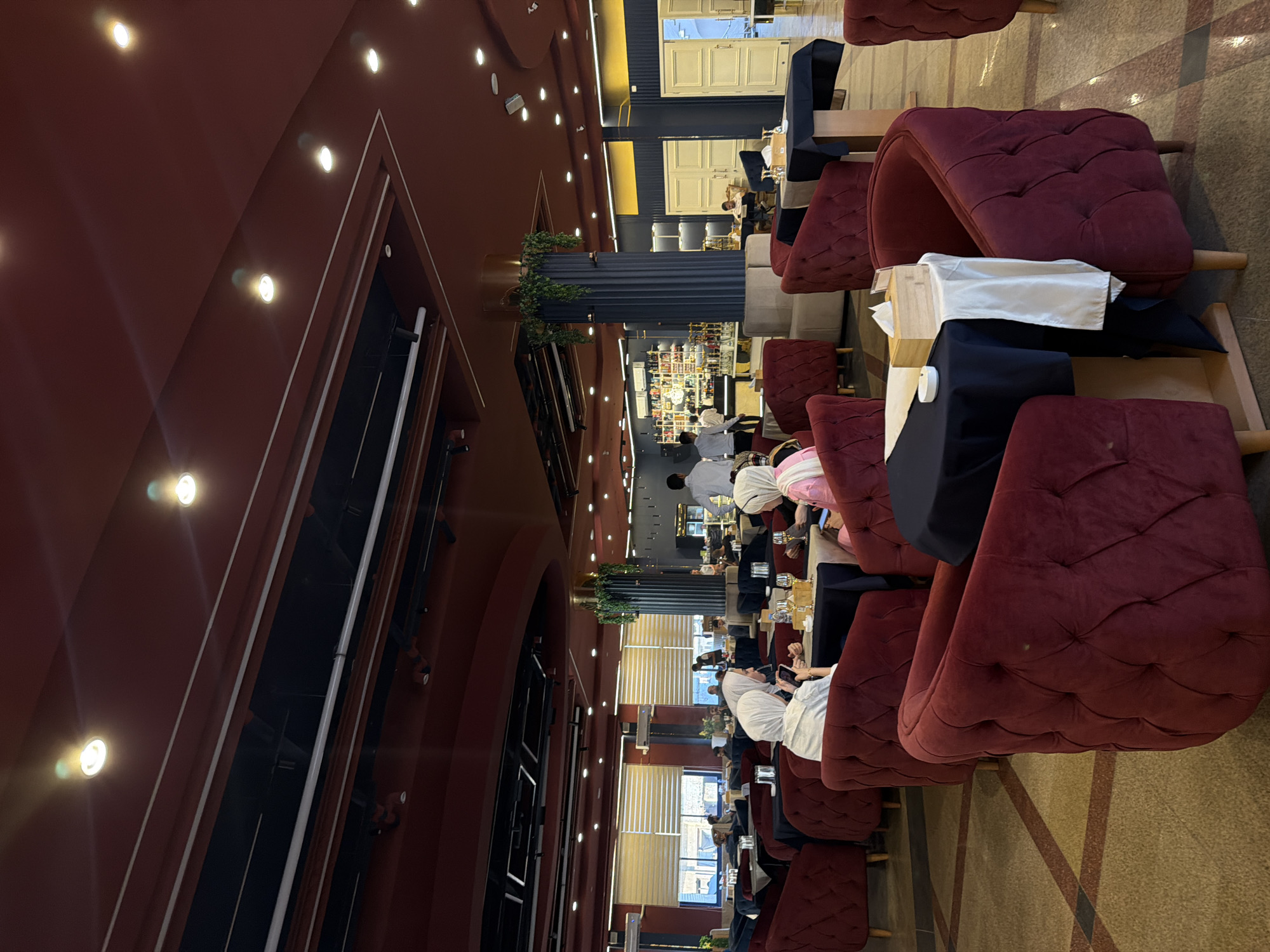 The image presents the interior of a spacious, modern restaurant or lounge, located in the city of Unknown, Unknown. The ceiling is primarily dark red, incorporating recessed circular spotlights that emit bright white light, with some exhibiting lens flare. Sections of the ceiling reveal exposed black structural elements and white pipes. The floor is covered with large, polished tiles in shades of beige, brown, and dark brown, arranged in a geometric pattern.
The furniture prominently features numerous dark red, tufted velvet armchairs with short wooden legs, positioned around square or rectangular tables. Most tables are covered with dark blue/black tablecloths, often accented with white fabric runners. On these tables, small wooden boxes (likely tissue dispensers) and white ashtrays are visible, alongside various drinking glasses, some containing beverages.
Approximately ten to fifteen individuals are present. In the mid-ground, a group of patrons is seated; at least two women are distinctly visible wearing white headscarves and light-colored tops, one engaging with a smartphone. In the background, several men, likely staff, are seen wearing white shirts. One male staff member is positioned behind a bar area, which displays an assortment of bottles and glassware. Another staff member is seen walking in the mid-left.
Architectural details include tall, fluted dark blue/black pillars adorned with hanging green plants. On the left side of the frame, large windows fitted with light-colored horizontal blinds are visible. On the right, several sets of light-colored, paneled double doors are present. Wall sections are painted dark blue/black, with some areas featuring yellow accents.