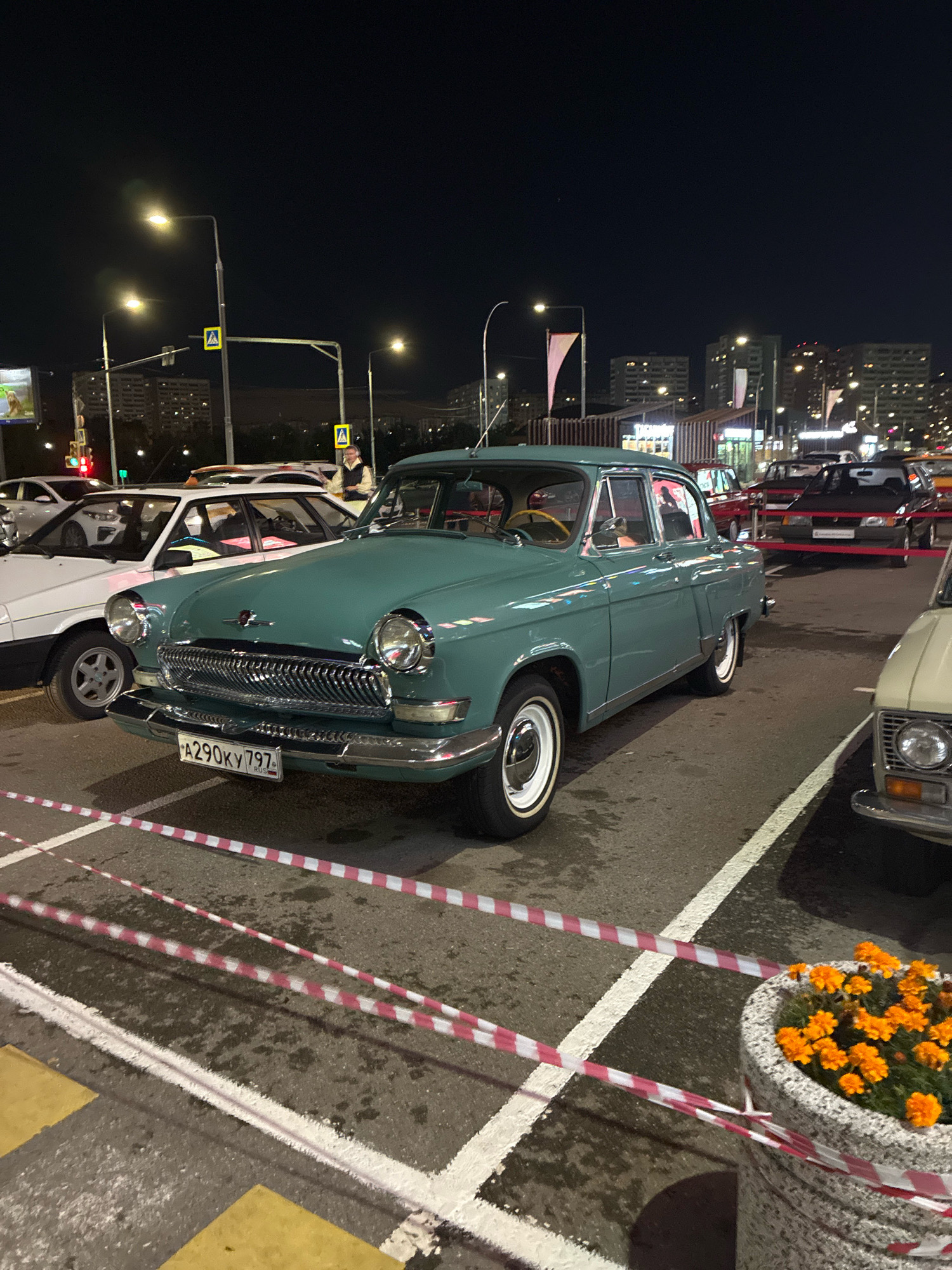 An outdoor, nighttime scene in Moskovskiy, Russia, features a gathering of vintage cars. In the foreground, a turquoise Volga GAZ-21 is parked diagonally in a lot, its chrome grille and white-walled tires catching the light. The license plate reads "A290KY 797". To its left, a white car, possibly a Lada, is partially visible. Behind the Volga, a red car and a dark-colored car are parked, separated by red tape. In the background, city buildings with illuminated windows are visible, along with streetlights and some flags. The ground is asphalt with white parking lines and some puddles. A decorative stone planter filled with orange marigolds is in the lower right corner, also cordoned off with red and white tape. No people are clearly identifiable as interacting with the cars.