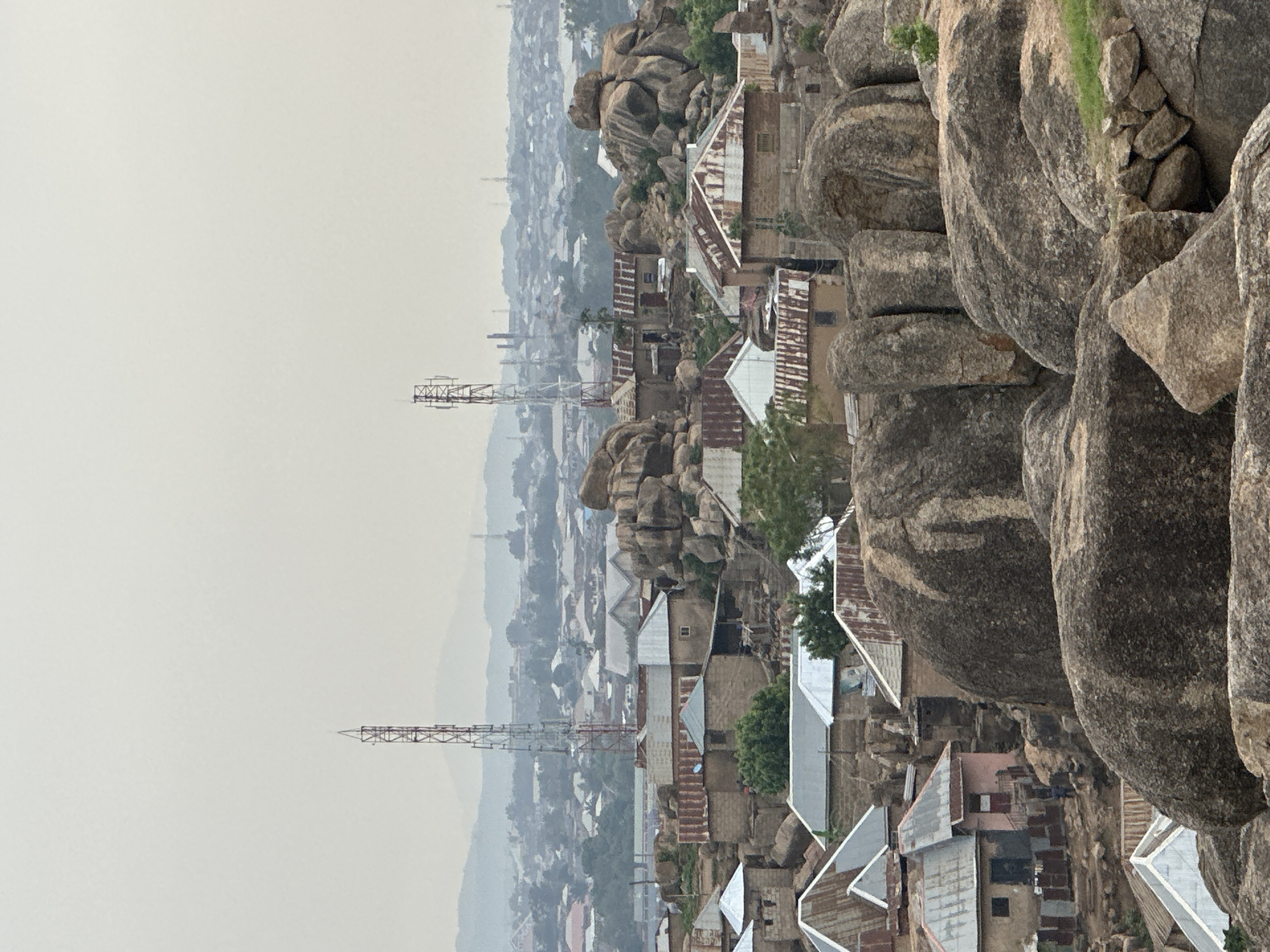 The image presents an expansive elevated view of a settlement intertwined with a rugged, rocky landscape, likely Jos, Nigeria, given the provided context. In the immediate foreground and mid-ground, large, smooth, grey-brown boulders dominate the scene, some with patches of sparse green vegetation. Nestled amongst these natural formations are numerous residential buildings, characterized by their varied architectural styles, including many structures with rusty or shiny corrugated iron roofs and walls constructed from blocks or bricks.
The settlement extends into the mid-distance, where a denser urban sprawl can be observed. Two prominent, tall lattice-style communication towers rise above the clustered rooftops, indicating modern infrastructure within the developing city. Beyond the immediate buildings, the city gradually fades into a soft, atmospheric haze, suggesting a considerable distance or an overcast, possibly dusty, weather condition. Faint outlines of distant hills or mountains are visible on the horizon, also softened by the atmospheric conditions.
The sky is a uniform, light grey, indicating an overcast day. The light is diffused, casting no strong shadows, which further supports the notion of an overcast or very hazy atmosphere. No explicit human activity is discernible, and the scene appears static, capturing a moment in time. There is no visible text in the image. The overall impression is one of human habitation adapting to and coexisting with a distinctive and challenging natural environment.