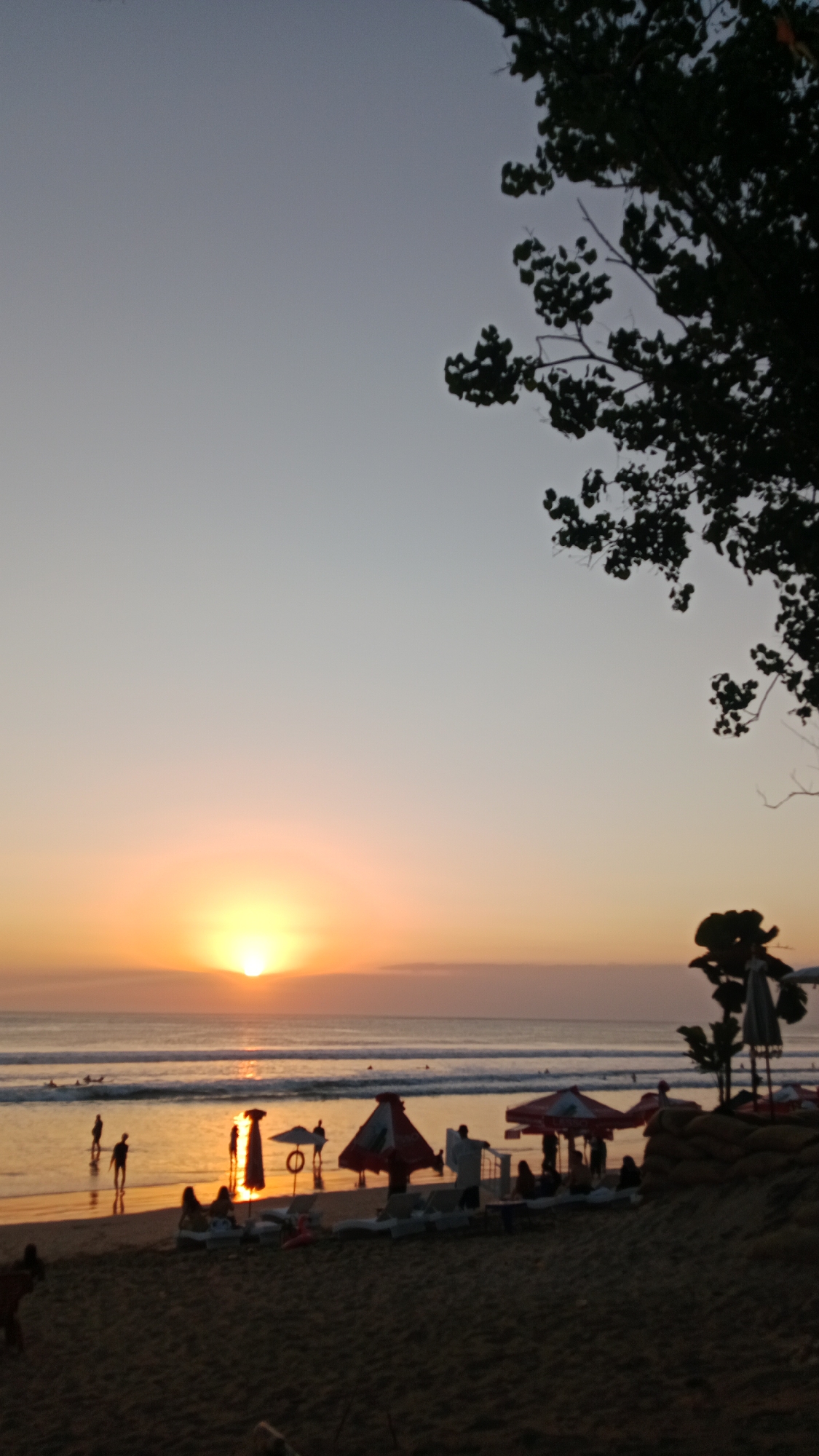 The image captures a sunset scene at Kuta Beach, Bali, Indonesia. The sun, a bright orb, is setting over the horizon, casting a warm, golden glow across the sky and the ocean. Gentle waves roll onto the sandy shore where several people are seen. Some are standing near the water, while others are relaxing on beach chairs, some of which are covered by umbrellas. In the foreground, a silhouetted tree branch hangs over the scene, adding a natural frame. The overall atmosphere is serene and peaceful, typical of a beach at dusk.