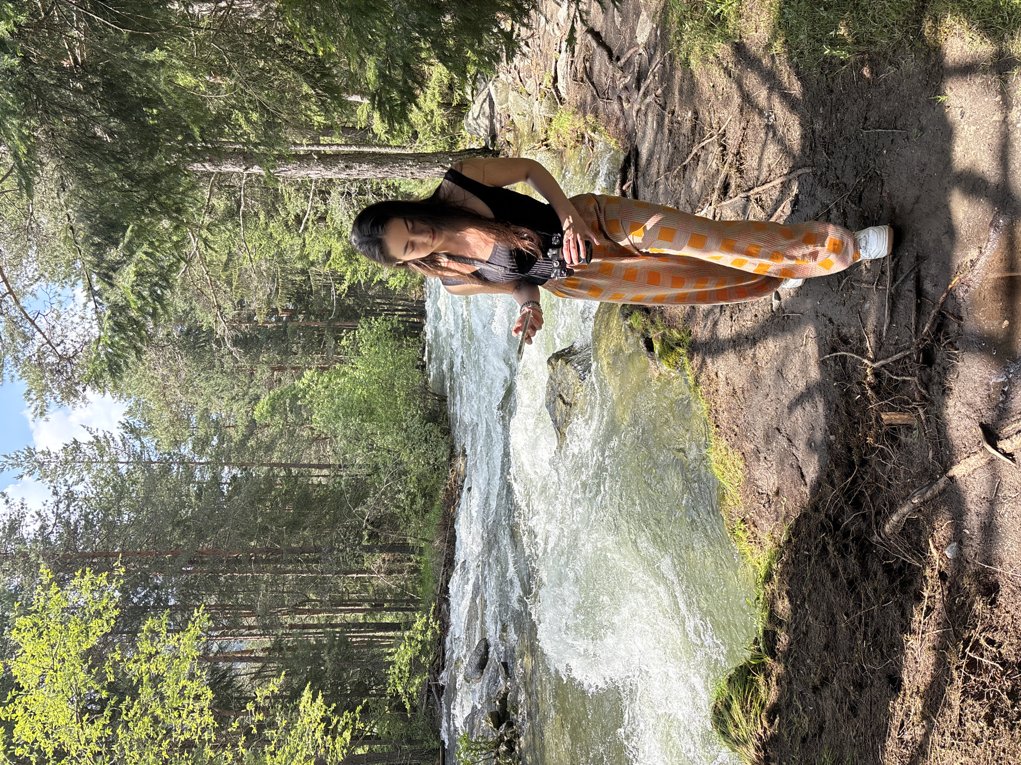 A woman is depicted walking along a dirt path beside a rapidly flowing river in a dense forest, specifically in the area of Beli Iskar, Bulgaria. The woman, with long dark hair, is dressed in a black sleeveless top and wide-leg trousers featuring a grid pattern of orange and lighter orange or beige squares. She wears white athletic shoes. In her left hand, she holds a silver-colored smartphone, while in her right hand, she carries a dark-colored camera, which has a strap draped around her neck. Her gaze is directed downwards towards the path as she walks.

The river, positioned to her left, exhibits significant turbulence with white rapids and visible rocks breaking the water's surface. The water has a light green tint in calmer areas. The path she walks on is unpaved, composed of dark earth with numerous exposed tree roots and scattered patches of green grass. Shadows from the surrounding trees and the woman herself are cast onto the ground, indicating bright overhead sunlight. To the woman's right, the forest continues with predominantly tall, slender coniferous trees and some deciduous foliage. The background features a dense growth of trees, suggesting a deep woodland environment. A small section of blue sky with sparse white clouds is visible through the tree canopy.