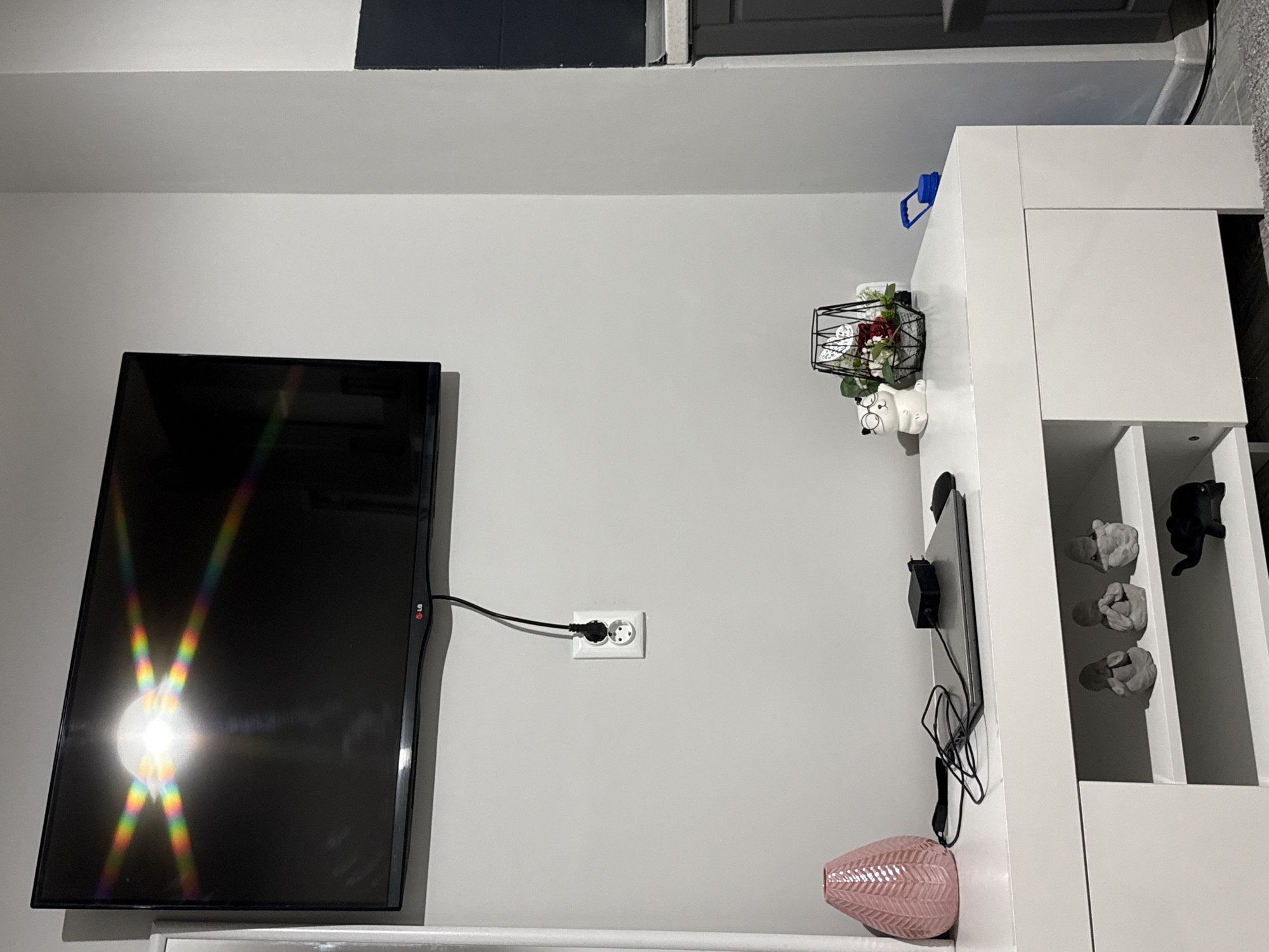 The image shows an interior wall with a television mounted on the left, plugged into an electrical outlet. To the right of the television, there is a white shelving unit with decorative items, including a flower arrangement, porcelain figures, and abstract sculptures. A pink vase sits on the top shelf. The room appears to be well-lit. The location is indicated to be in Stara Zagora, Bulgaria.