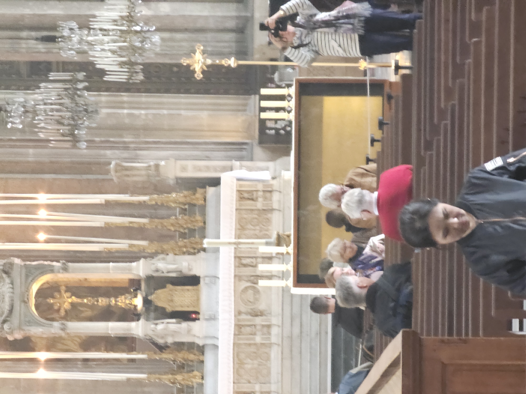 The image shows a section of the Notre Dame Cathedral in Paris, France. A group of tourists is viewing the altar area. One person is taking photos, and others are standing or sitting in the pews. Candles are lit near the altar, and chandeliers hang from the ceiling. The architecture is visible, including the stone work and details of the altar and the pews.