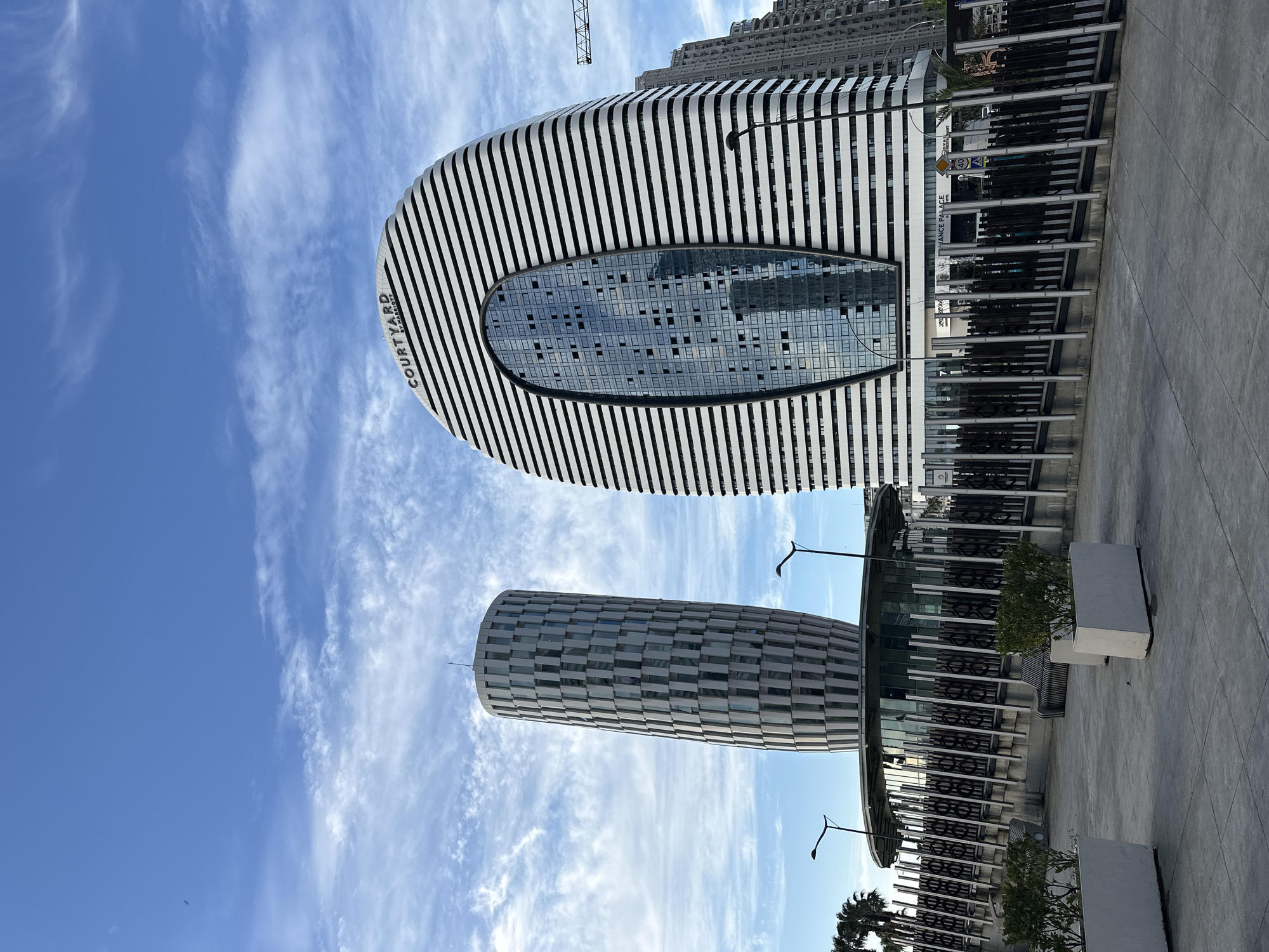 The image shows two prominent buildings in Batumi, Georgia, against a backdrop of a partially cloudy blue sky. The first building is the Courtyard by Marriott hotel, characterized by its curved shape and black and white striped facade. The center of the building features a large oval-shaped glass section, reflecting the surrounding sky and cityscape. The hotel's name, "COURTYARD BY MARRIOTT," is vertically displayed on the building's side. The second building, also with a curved design, is covered in a checkerboard pattern of gray and white. Both buildings are situated near a public area or plaza, marked by a gray concrete surface, black metal barriers, and greenery in planters. In the background, taller buildings and a crane are visible, indicating the urban environment. The scene is photographed from a low angle, emphasizing the height and architectural features of the buildings. The image appears to be taken during daylight hours.
