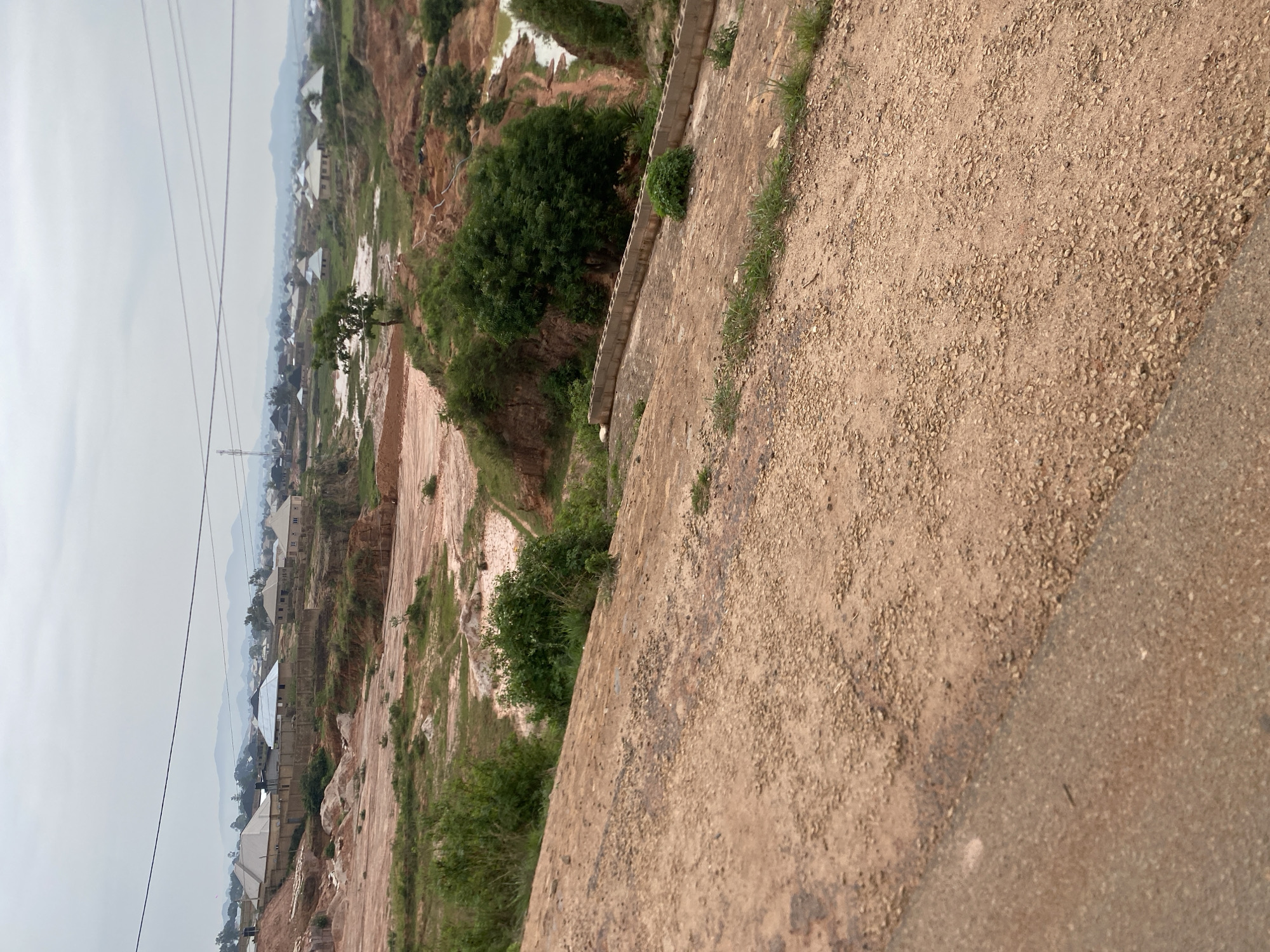 The image captures a landscape in Reifild-Ist, Nigeria, characterized by a rural or semi-developed setting. The foreground is dominated by a dirt and gravel surface, possibly a road or path, with scattered small stones and patches of dry grass. In the midground, a sloped terrain reveals reddish-brown soil, indicating possible erosion or construction activity. Various low-lying bushes and trees are interspersed across this area, with some larger trees contributing to the greenery.
Further into the scene, a man-made structure, a low wall constructed from concrete blocks, runs horizontally across the terrain. Beyond this, the landscape descends into a valley with muddy water, suggesting recent rainfall or a water source. The valley walls are steep and eroded, showing layers of soil.
In the background, a settlement of houses with distinct rooflines, some conical and others angular, is visible on elevated ground. The houses appear to be of modest construction, with some enclosed by fences. The distant horizon is marked by rolling hills and mountains, softened by atmospheric haze. Overhead, power lines crisscross the sky, indicating the presence of electrical infrastructure. The overall atmosphere is subdued, with an overcast sky contributing to a diffused light. There are no people or animals visible in the image.
