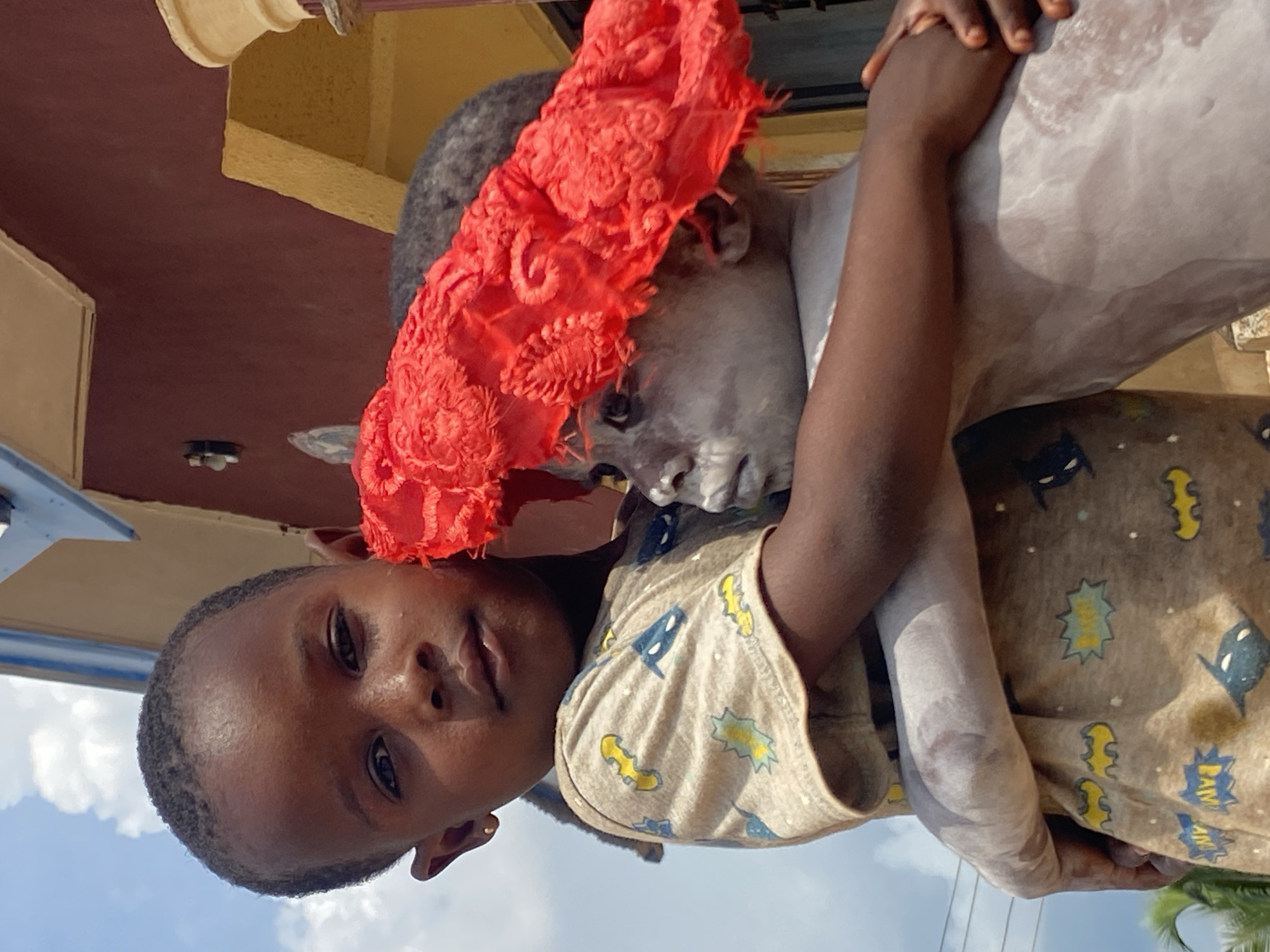 Two young Black children are hugging each other in Amoso, Nigeria. One child, facing the camera, wears a beige t-shirt with cartoon motifs. The other child, partly covered in white powder, has a red ornamental headdress. The scene is outdoors, with the sky visible at the bottom of the frame and a building partially seen in the background.