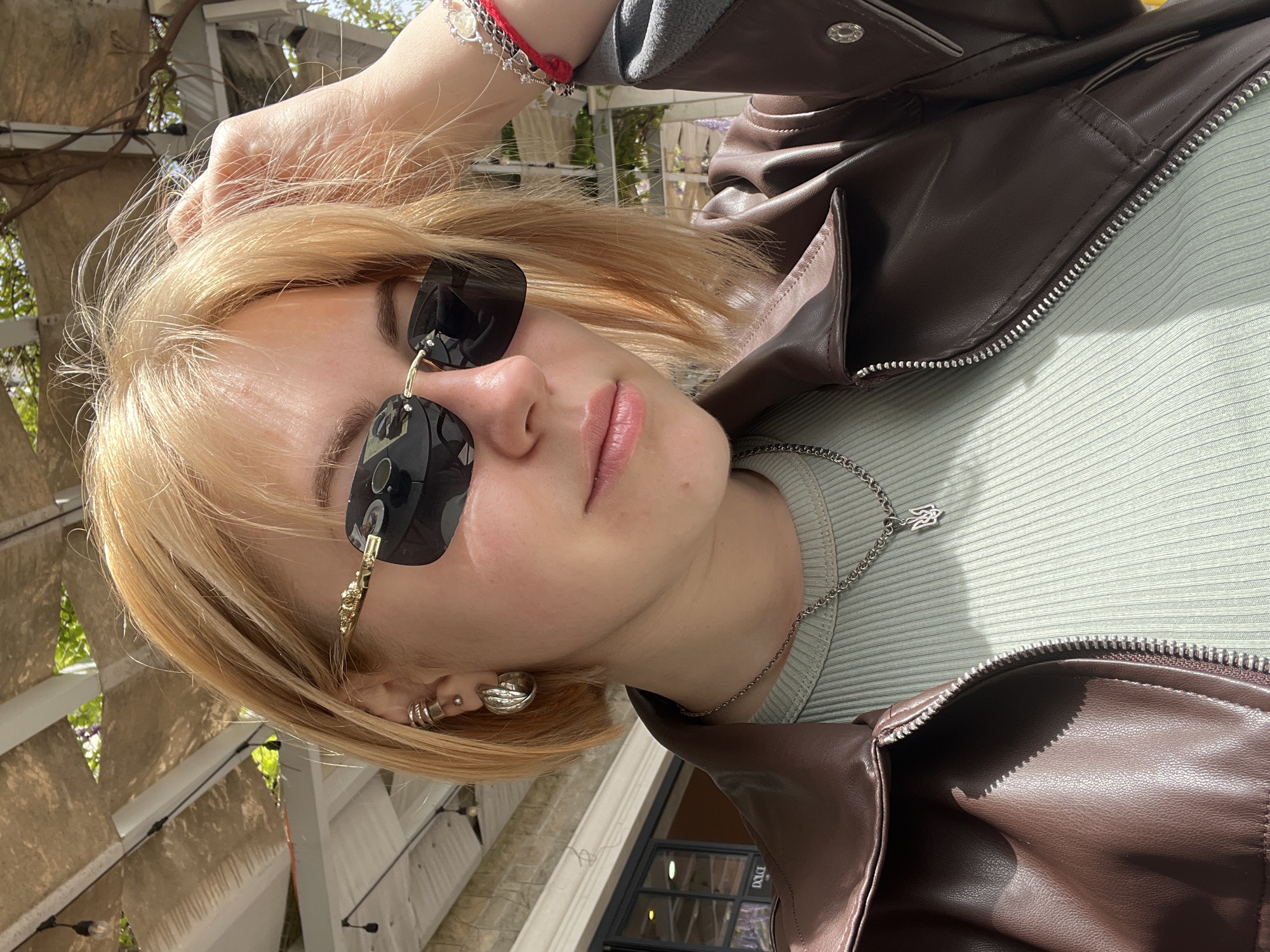 A young woman with blonde hair poses outdoors. She is wearing sunglasses, a brown leather jacket over a light green ribbed top, and jewelry including a silver necklace and earrings. She is holding her hair with her hand. The location appears to be Rafailovići, Montenegro, based on elements visible in the background: a stone structure, wooden beams, and foliage that are consistent with the architecture and vegetation typically found in that coastal area. The photo seems to be taken during the day.