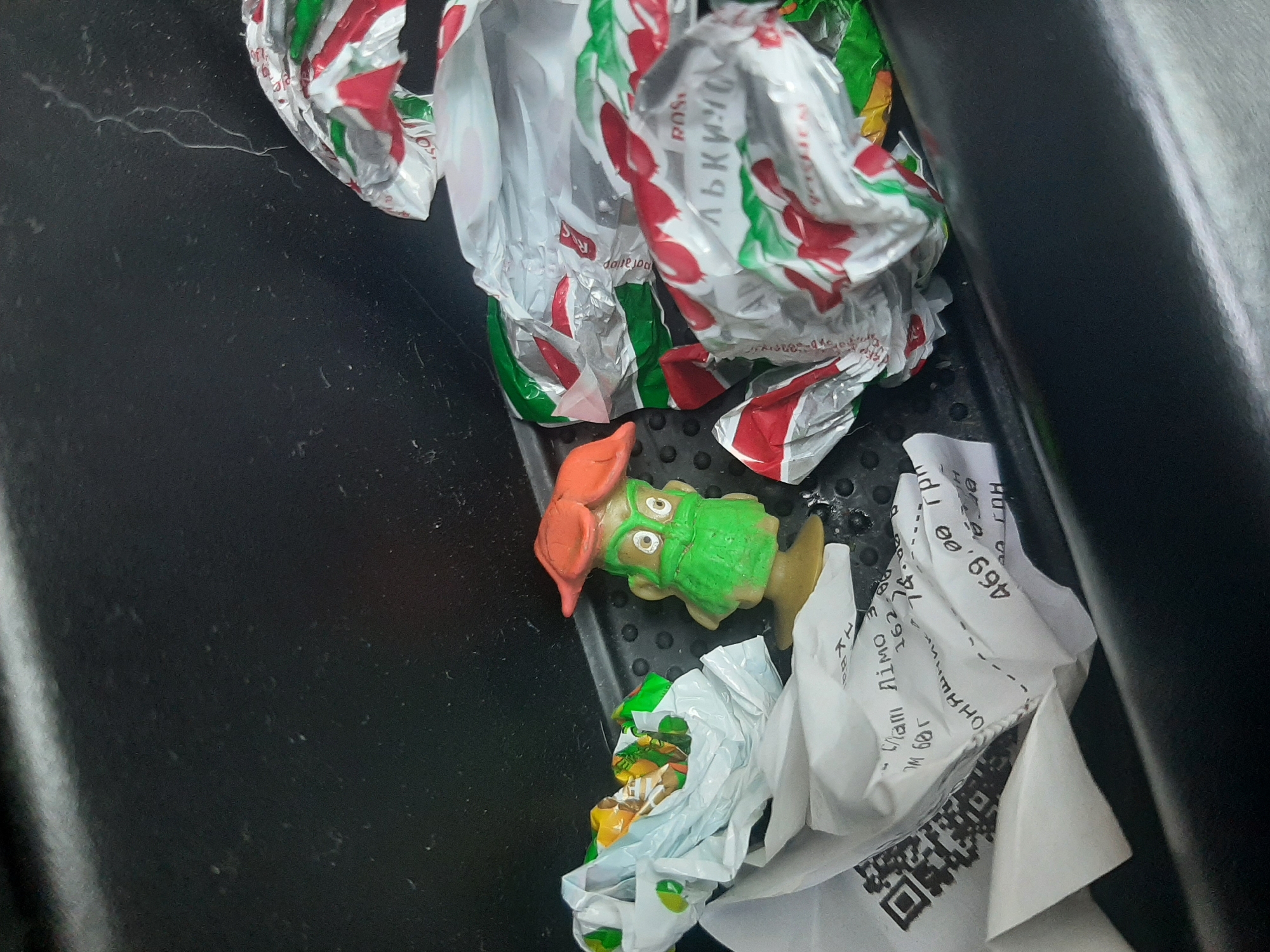 A small green plastic toy figure with orange hair and large eyes stands on a light brown base. It is positioned in the center of the frame, slightly to the left. To the left of the toy, there is a crumpled white plastic bag filled with small, colorful objects, possibly candy wrappers. To the right of the toy, there are several crumpled wrappers with red and green patterns, also appearing to be candy wrappers. Below the toy and to its left, a white receipt is visible, partially crumpled and bearing text and a QR code. The background is a dark, textured surface, possibly the interior of a car or a storage compartment. The lighting is dim, with a bright light source coming from the top left of the frame, casting shadows.