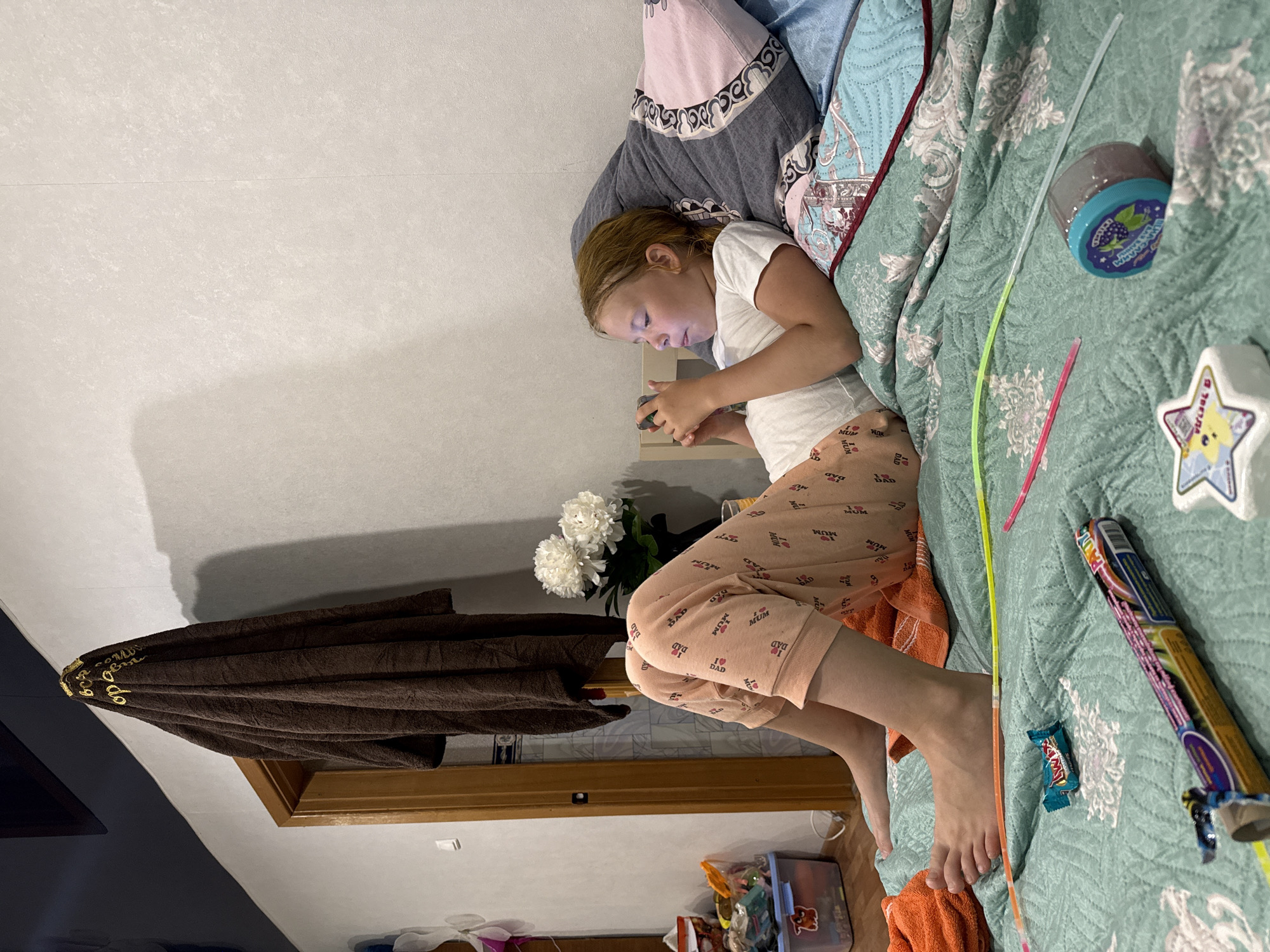 A young girl is lying on her side on a bed, looking at her phone. She is wearing a white t-shirt and light pink pajama pants with a pattern of hearts, "MUM", and "DAD" on them. The bed has a teal patterned comforter, and there are pillows with different designs. Scattered on the bed are glow sticks, a jar of purple slime, and what appears to be a candy wrapper. To the left of the bed, a brown towel is draped over a wooden structure, and a bouquet of white flowers sits near the edge of the bed. The overall scene suggests a relaxed, indoor setting, possibly a bedroom or a living room. The lighting suggests it could be daytime, but the absence of windows makes it difficult to determine the exact time of day or weather. The girl's focus on her phone indicates she is engaged in a digital activity. There is visible text on her pajama pants, including "MUM" and "DAD" within heart shapes, and a star-shaped object with Russian text that translates to "I am a star."