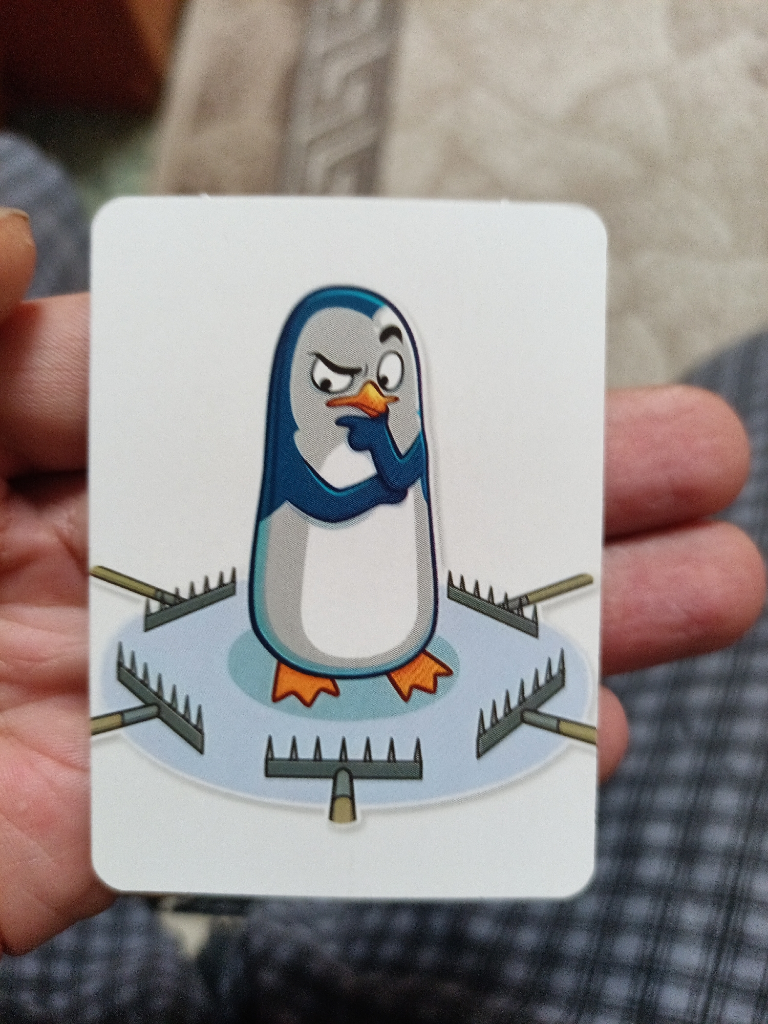 An image shows a card with a cartoon penguin character standing on a circular patch of ice. The penguin appears to be deep in thought, with one flipper raised to its beak and an eyebrow furrowed. Around the penguin, on the ice, are several garden rakes, arranged in a circle. The rakes are positioned with their tines facing upwards. The card is held in a person's hand, with parts of the fingers and thumb visible. The background is blurred, showing a textured surface. There is no discernible location in the city of Nyagan, Russia.