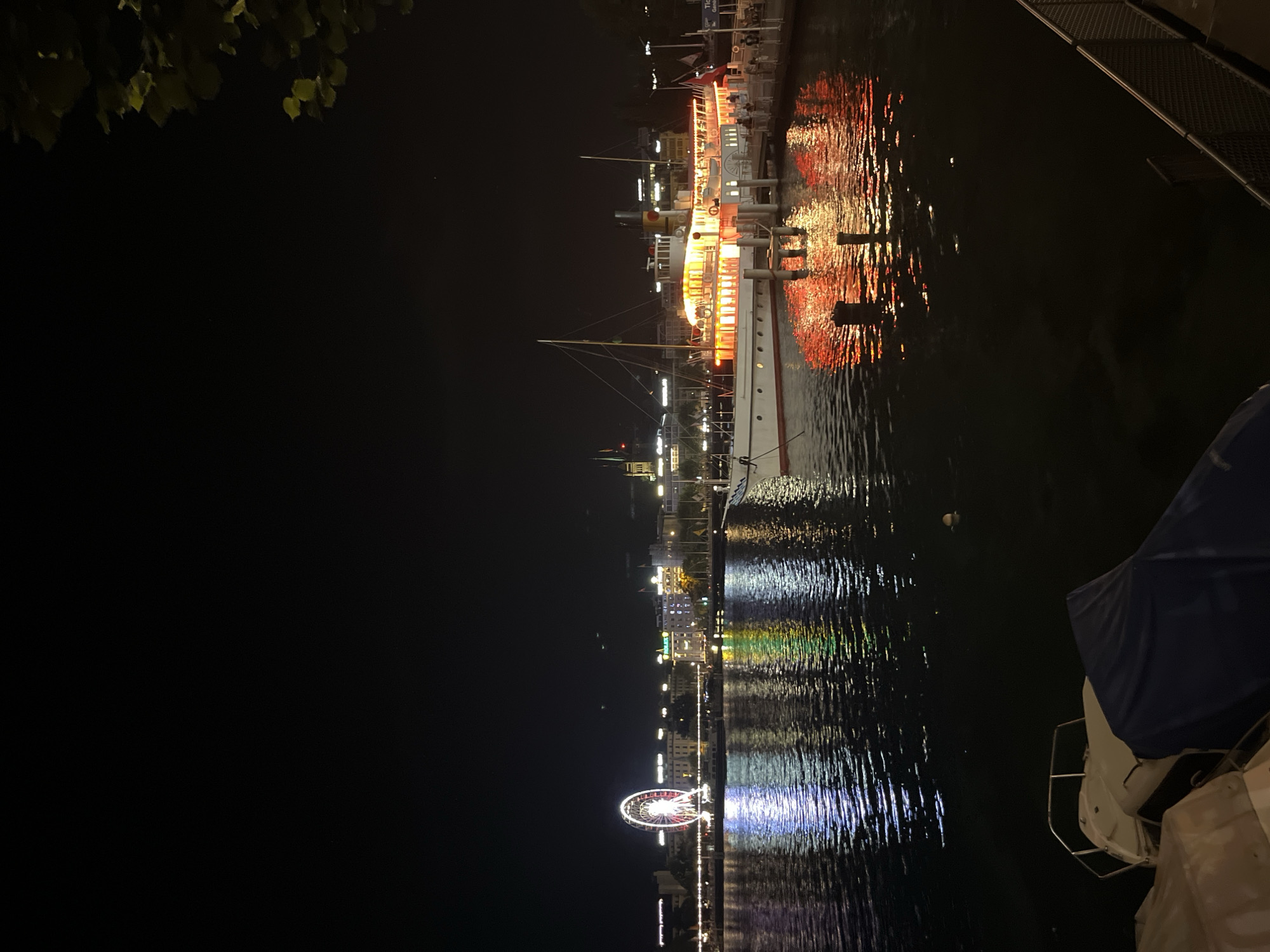 This is a nighttime, eye-level shot of the Jet d'Eau promenade in Geneva, Switzerland, looking across Lake Geneva towards the city skyline. The image captures the reflection of the city lights on the water. Prominently featured in the foreground is a docked vintage steamboat, its windows illuminated with warm orange light, casting vibrant reflections onto the dark water. In the mid-ground, the city skyline stretches across the horizon, dotted with illuminated buildings and a large Ferris wheel on the left, also lit with colorful lights. The water is dark and rippling, reflecting the lights from the city and the steamboat. In the lower left corner, the sterns of two covered boats are visible. A dark fence runs along the right edge of the promenade. A few individuals are visible on the dock near the steamboat, and a couple of buoys are floating in the water.