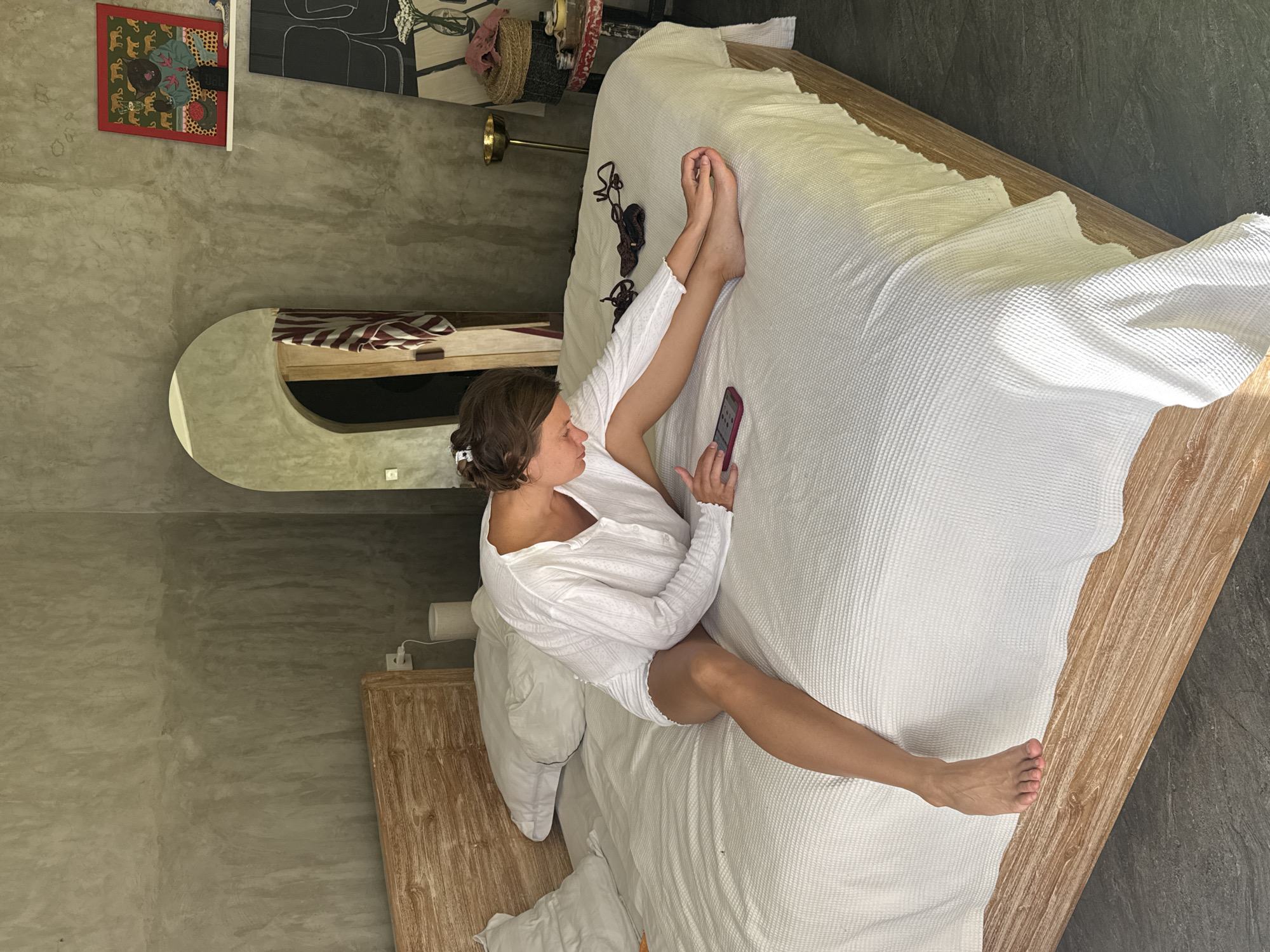 A woman is lying on a bed and using a smartphone. She is wearing a white, long-sleeved top and shorts. Her feet are extending off the end of the bed. The bed is white with a wooden frame and headboard. A nightstand is visible on the left side of the bed, as is a mirror and some artwork on the wall behind the bed. The room appears to be a bedroom with concrete walls and flooring.
The location is North Kuta, Indonesia.