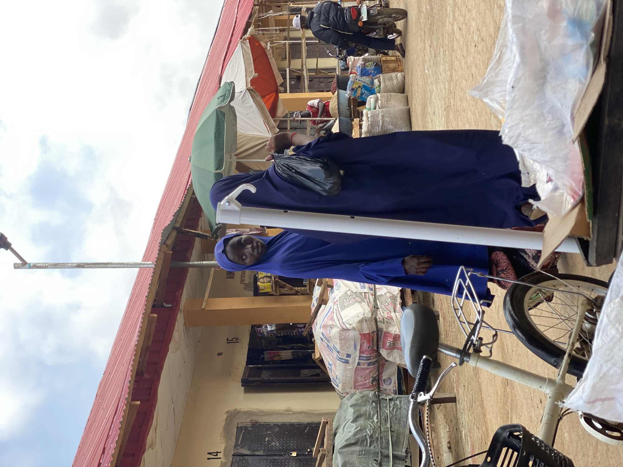 A person in a blue hijab stands near a white bicycle and a building labeled "14" and "15" in what appears to be a marketplace in Kafanchan B, Nigeria. Other individuals are visible in the background, along with motorcycles, tents, and various goods typically found in a market setting. Bags of goods, possibly food items, are stacked near the bicycle.