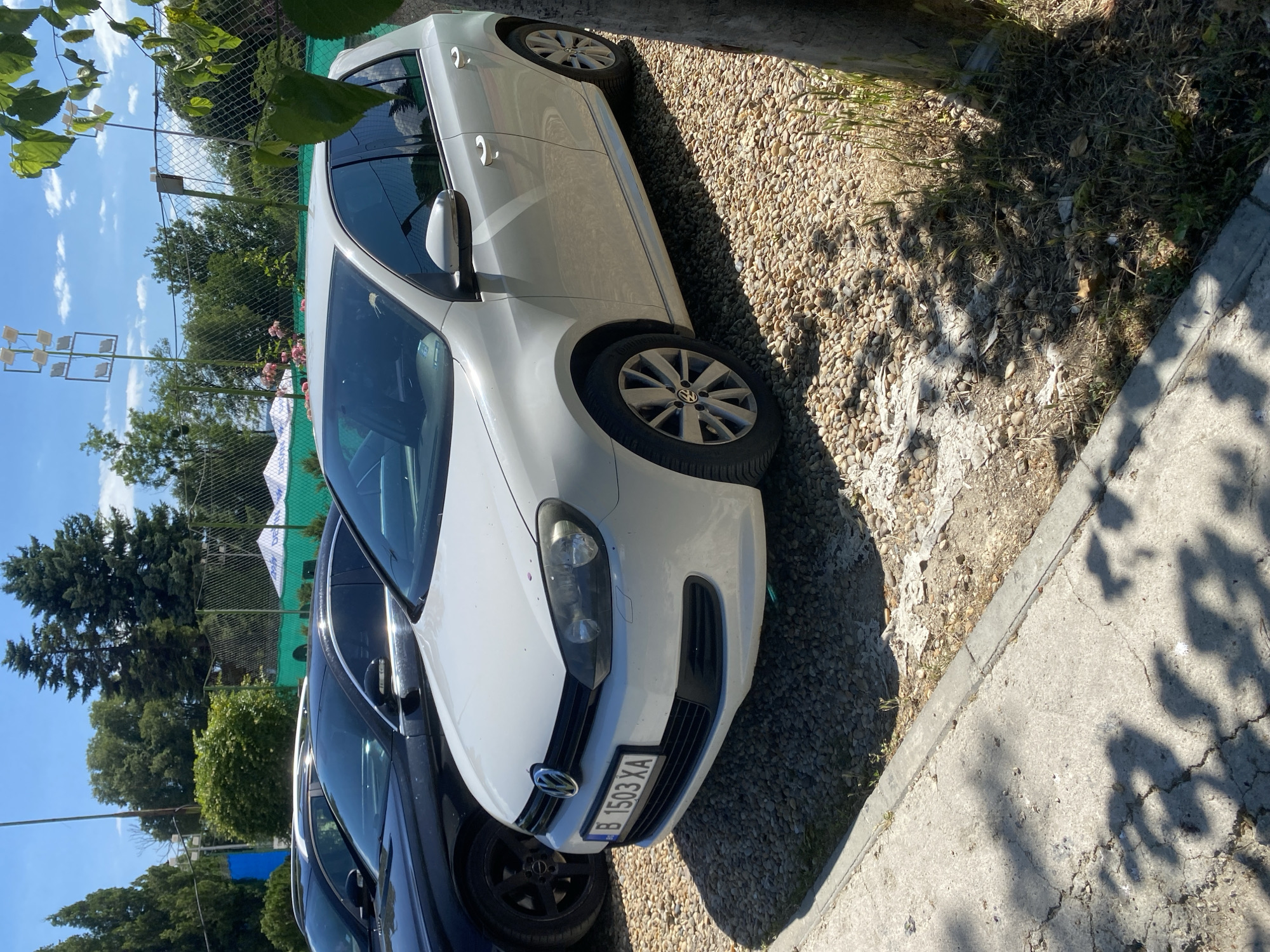 A white Volkswagen Golf Mk6 is parked on a gravel lot. The car is facing right, with its front bumper visible. The license plate reads "B 1503 XA." In the background, there is a green fence and trees. The sky is blue with some clouds. The scene appears to be outdoors in a sunny location. The car is likely in the city of Varna, Bulgaria, given the license plate format.