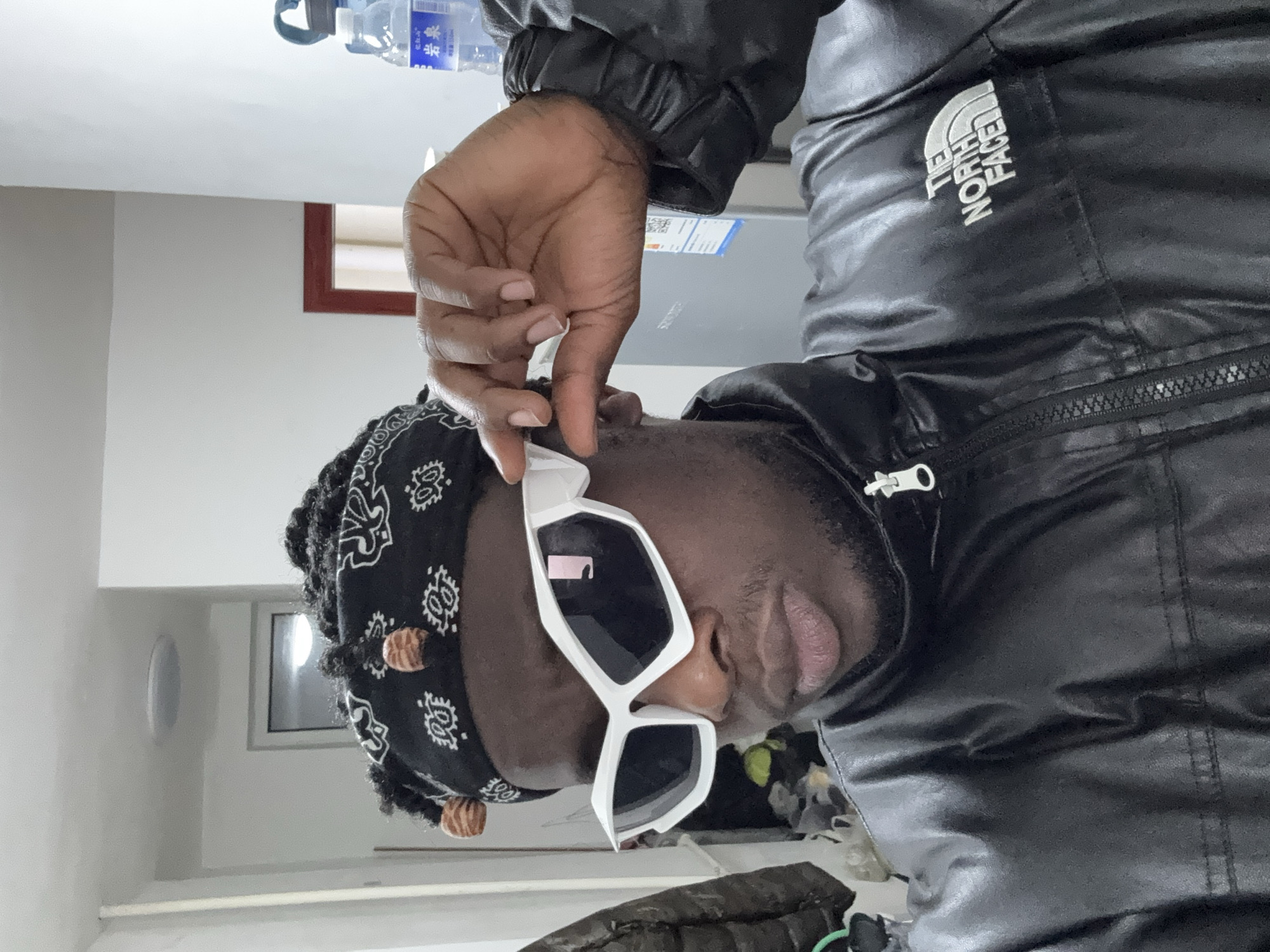 A person with dark skin is depicted indoors, wearing a black bandana with white patterns and decorative cylindrical beads. The person is also wearing white, angular sunglasses. A black, shiny jacket with "THE NORTH FACE" logo is visible. The person is holding the sunglasses with their right hand. Background elements include a white wall, a window, a water bottle, and a portion of what appears to be a refrigerator. The exact location within Zhongtun, China is not discernible from the given image.
