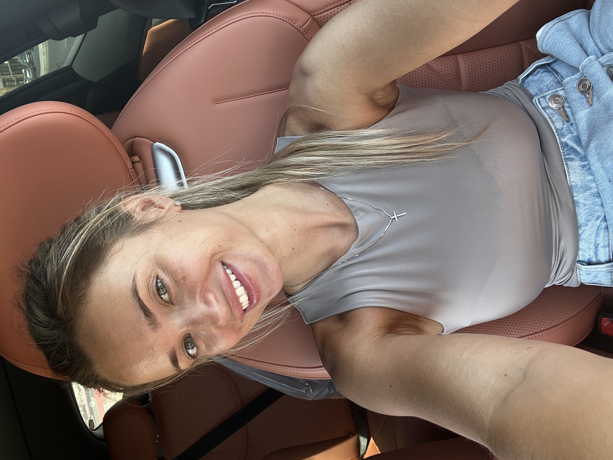 A woman is sitting in the driver's seat of a car. She has blonde hair pulled back in a ponytail and is smiling at the camera. She is wearing a grey tank top and blue jeans. The car has brown leather seats. The image appears to be taken outdoors, possibly in a parking lot.