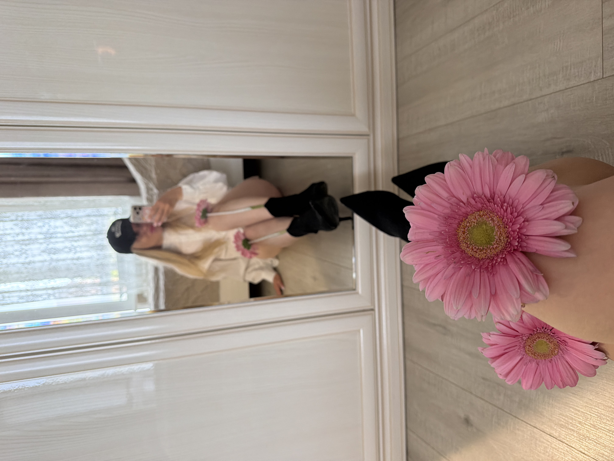 The image shows a selfie reflected in a mirror. The person in the reflection is holding a phone, taking the picture. They are wearing a light-colored top, black leggings, and black heeled boots. They have a black baseball cap on. In the foreground, two pink gerbera daisies are visible, partially obscuring the scene. The background includes a white paneled door or wall and a light-colored, plank-patterned wall. There is no evidence to indicate this image was taken in Votkinsk, Russia.