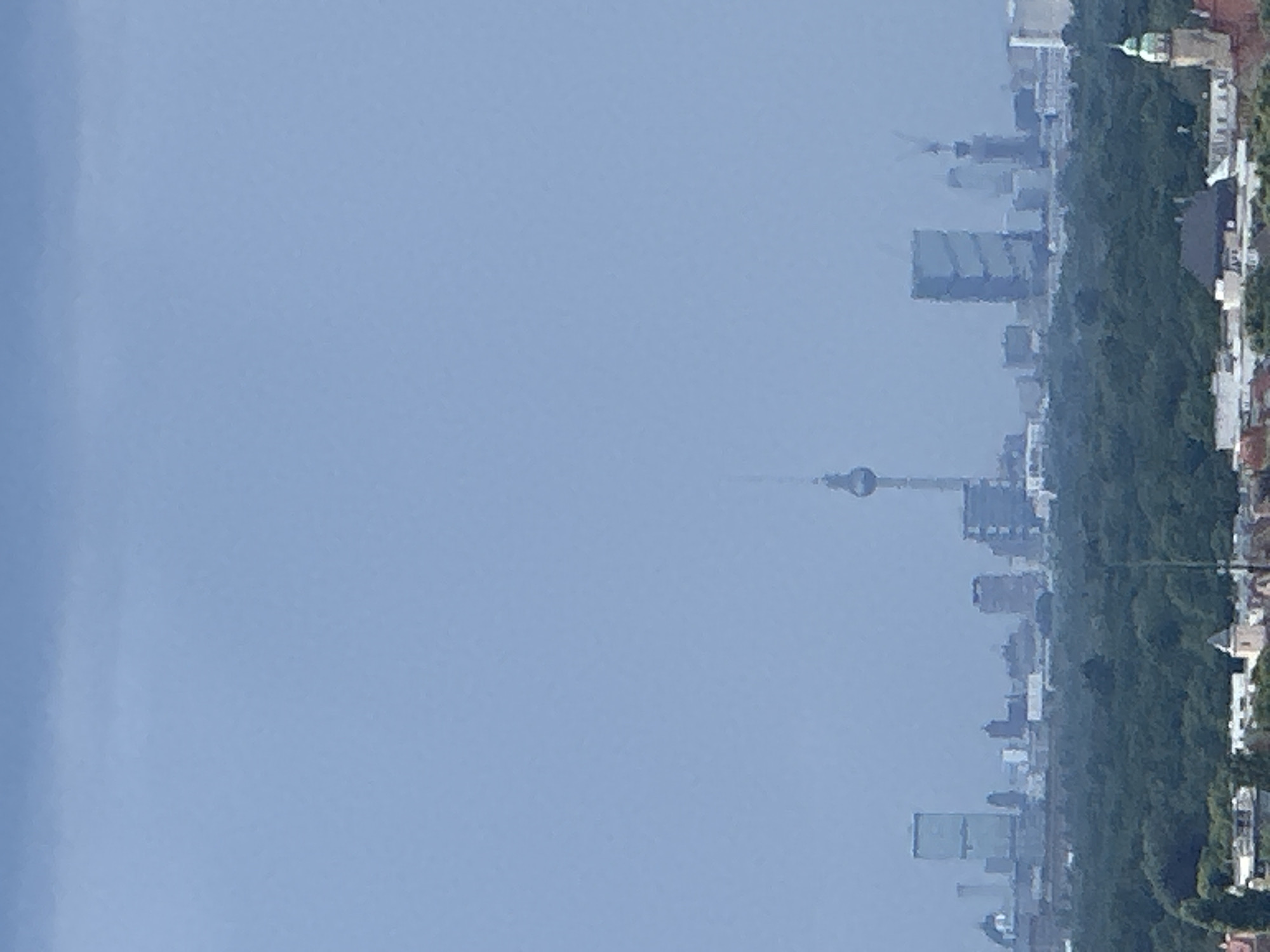 The image captures a hazy panoramic view of the Berlin skyline, dominated by a muted, overcast sky. In the foreground, a dense, dark green forest stretches across the lower portion of the frame, providing a natural buffer between the viewer and the urban landscape beyond. Above the treeline, a collection of buildings comprises the city's silhouette.
Prominently positioned in the middle ground is the Fernsehturm (Television Tower), a slender, cylindrical structure topped with a spherical observation deck and antenna, standing tall against the hazy backdrop. To its left, a light-blue, rectangular skyscraper with distinct vertical lines stands out. To the right of the Fernsehturm, a cluster of modern buildings includes a taller, dark blue skyscraper and a building with visible construction cranes. Various other buildings of varying heights and architectural styles are scattered throughout the skyline, some appearing as indistinct shapes due to the atmospheric conditions.
The lower edge of the image reveals the tops of some buildings and structures, suggesting a lower elevation closer to the viewer. A small, light-colored tower with a green dome is visible on the far right, hinting at older architecture amidst the modern structures. The overall impression is one of a vast, somewhat muted cityscape viewed from a distance, with the weather contributing to a subdued color palette and reduced visual clarity.