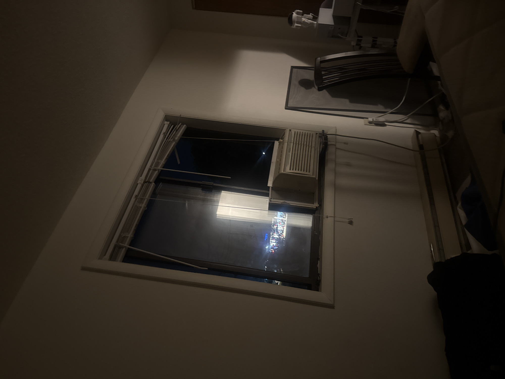 This is an indoor, eye-level, medium shot of a room at night. The room appears to be a bedroom, with a bed in the foreground covered by a dark blanket. To the left of the bed is a dark object, possibly clothing. In the background, a window shows a dimly lit cityscape, suggesting the location is the City of Binghamton, United States of America. The window has blinds that are partially open, and an air conditioner is installed in the lower part of the window frame. To the right of the window, there is a chair, a framed object leaning against the wall, and some medical equipment on a stand. The walls are painted white, and the lighting in the room is dim, with shadows cast by the objects.