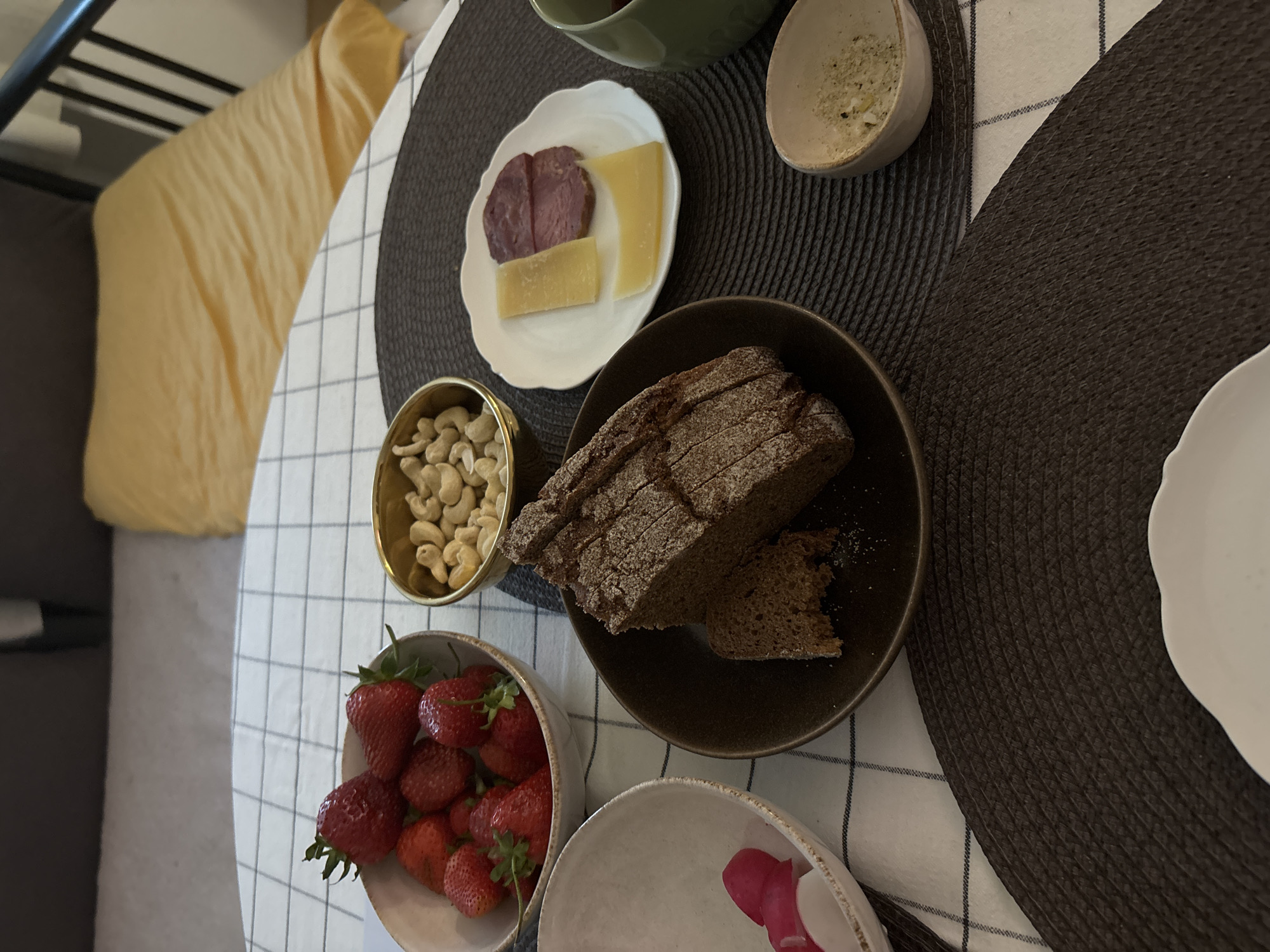The image depicts a table setting, possibly indoors, with various food items arranged on a checked tablecloth. Several bowls and plates contain strawberries, cashews, sliced bread, cheese and salami, and a green bowl. The arrangement suggests a meal or snack setting. The exact location cannot be determined from the image. There is no indication of the city of Saint Petersburg, Russia. No people are visible.
