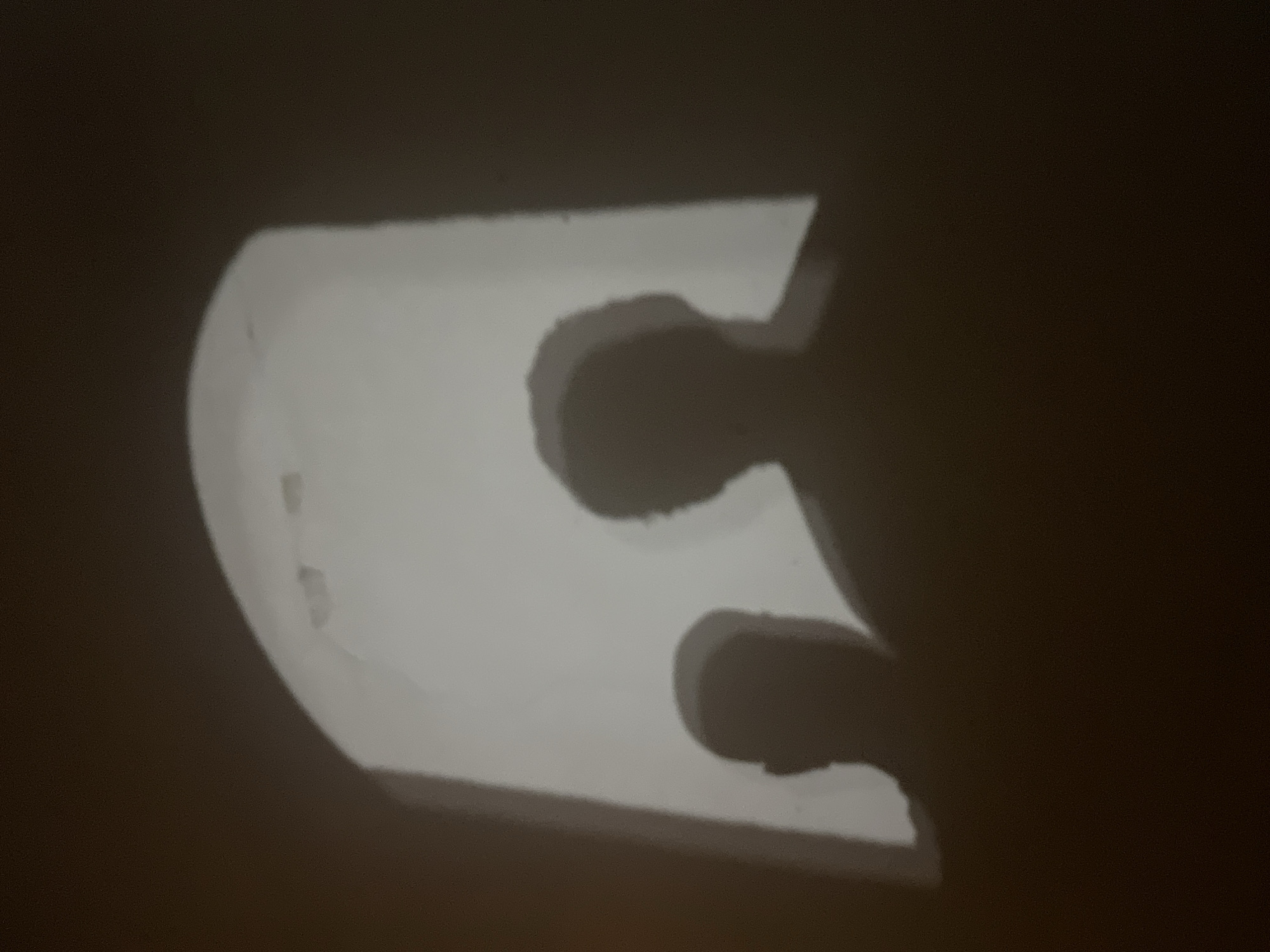 A brightly lit, white plastic part, possibly a clip or fitting, casts a shadow on a dark surface. The object has a rounded edge on the left and a more angular shape on the right, with two holes visible through the material that also cast shadows. The overall lighting suggests a single light source illuminating the object against a dark background. There is no information in the image to indicate the location in Budva, Montenegro.