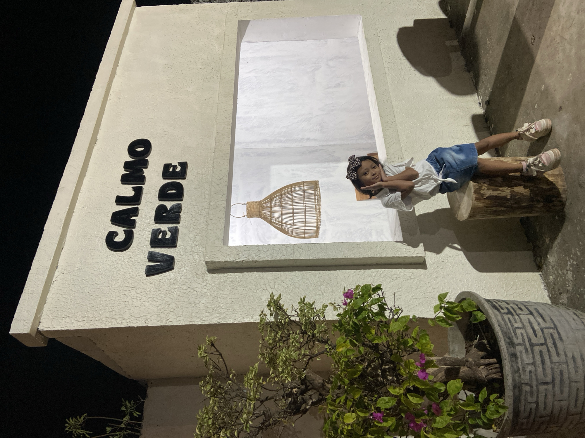 A young girl with a black and white patterned headband sits on a wooden log in front of a textured white building with "CALMO VERDE" in black 3D lettering above a rectangular window. The girl wears a white knotted shirt, a denim skirt, and light-colored sneakers. Her hands are placed on her cheeks, and she is looking at the camera. To the left of the building, a large potted plant with green leaves and purple flowers is visible. The scene is illuminated by artificial light, casting shadows on the ground. The background is dark, suggesting it is nighttime. The location is the city of Kesiman, Indonesia.