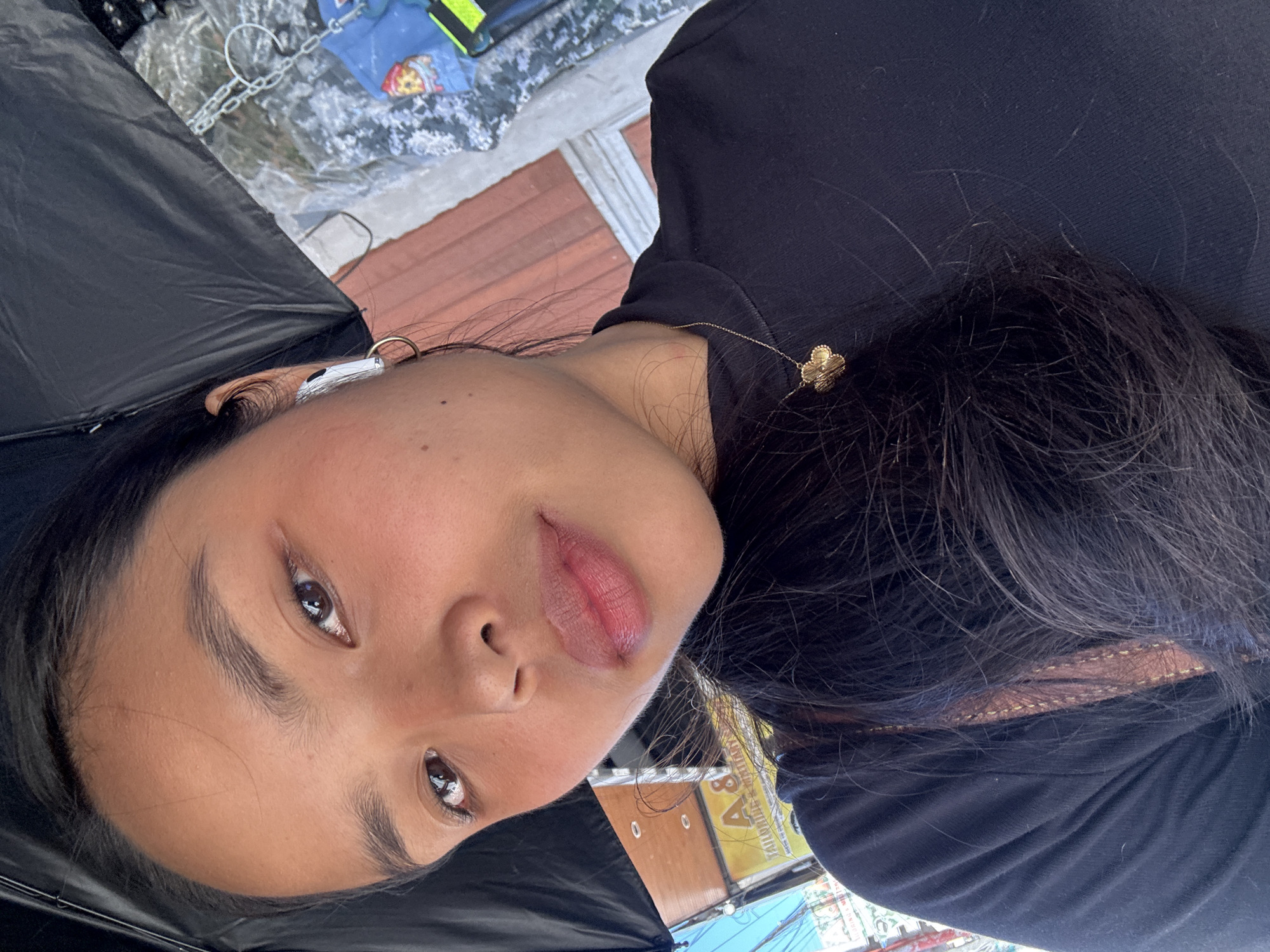 A close-up selfie shows a young woman holding a black umbrella, partially obscuring the upper portion of the frame. She is wearing a black top and a gold necklace with a clover-shaped pendant. She has dark hair, brown eyes, and is wearing an earbud in her left ear, along with a gold hoop earring. The background suggests an outdoor urban environment in Davao City, Philippines, with hints of a building facade, signage that appears to read "A & S Tailoring & Embroidery," and other elements typical of a street scene. The lighting suggests it is daytime.