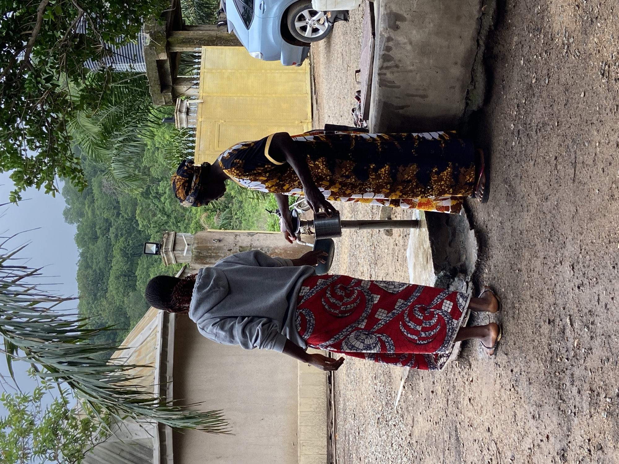 The image captures an outdoor scene in Mararaba, Nigeria, featuring two individuals near a communal water tap.
On the left, a person is observed from behind, wearing a grey hooded sweatshirt and a long red skirt patterned with white and black swirling motifs. They wear light brown flat sandals and stand on unpaved ground consisting of dirt and small gravel.
To the right, a second person is viewed in left profile, wearing a multi-colored headwrap and a long, loose-fitting dress adorned with black, yellow, and white patterns. This individual is actively operating a metal water tap, filling a silver metal container with water. Dark flip-flops are visible on their feet. The water tap is affixed to a vertical metal pipe emerging from a concrete base, which is visibly wet from water overflow.
The background is dominated by dense green foliage and numerous trees, indicating a lush, possibly forested environment. Above the vegetation, the sky is uniformly overcast and grey. Architectural elements include a building on the left with a rusted corrugated metal roof. To the right, a tall concrete structure supports a striped cylindrical object, likely a water tank. Below this, a large yellow metal gate with decorative elements and a small integrated door is visible. An outdoor floodlight is mounted on a concrete pillar near the gate. A light blue SUV is partially visible parked behind the gate on the far right. A prominent palm frond extends into the upper left portion of the frame. The ground throughout the scene is unpaved, a mixture of damp earth and stones.