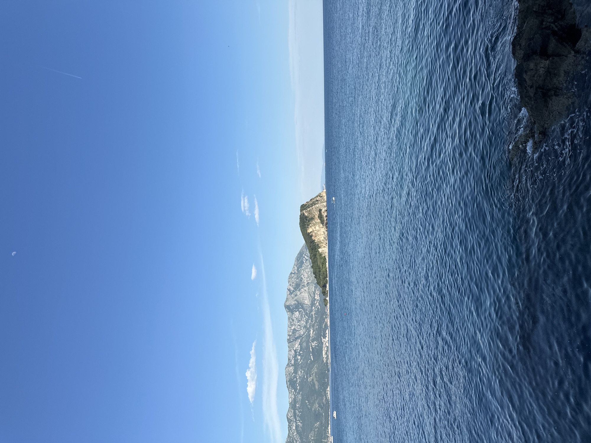 The image shows a coastal scene, possibly near Budva, Montenegro, featuring a clear blue sky taking up the left half of the image, contrasting with the deep blue sea on the right. A rocky cliff, displaying geological formations and some vegetation, separates the sky from the water. The sea's surface reveals textured patterns of small waves. There is a hint of an airplane trail in the sky. The lighting suggests a sunny day.