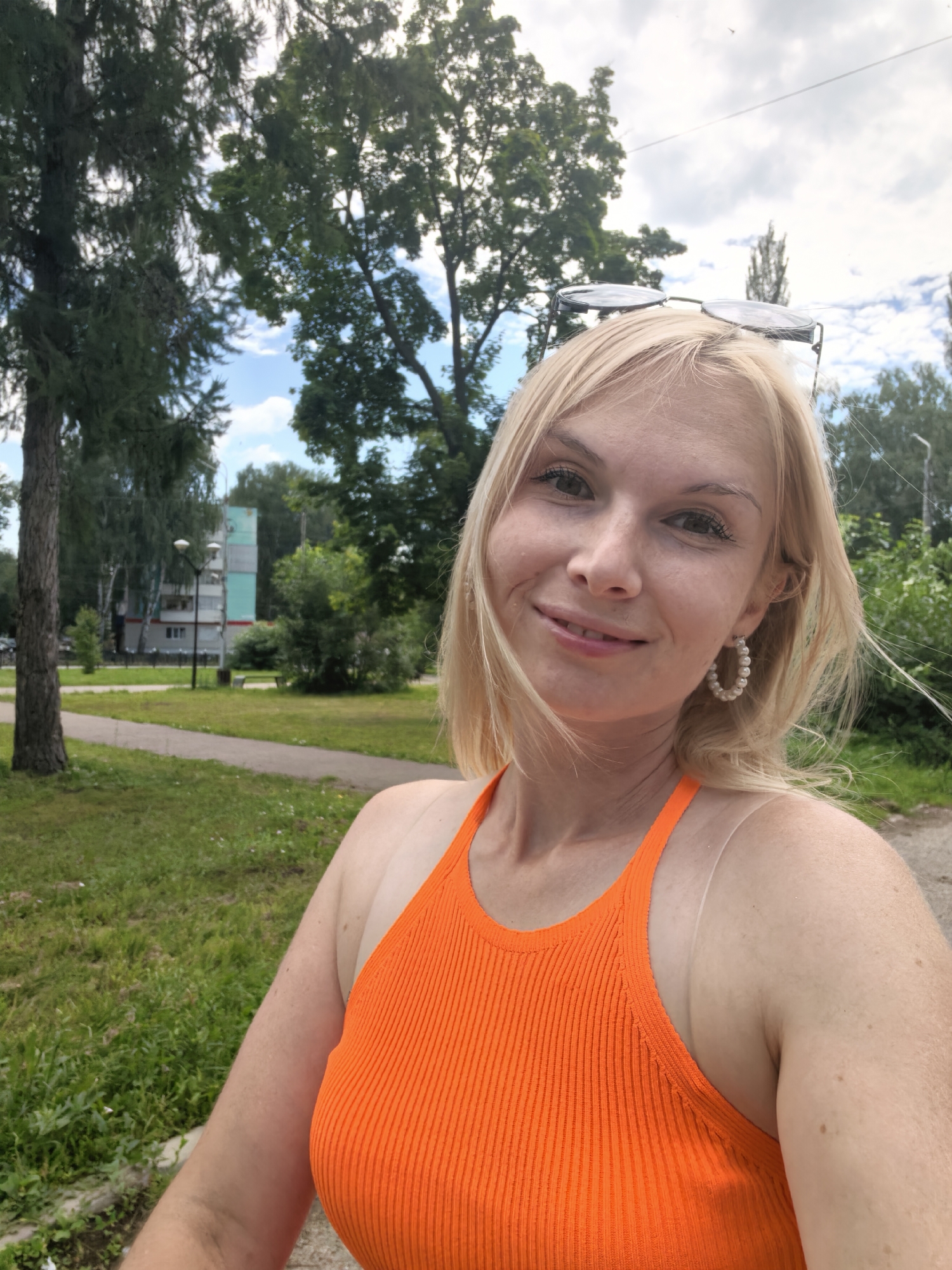 A blonde Caucasian woman is taking a selfie outdoors in a park-like setting with trees and grass. She is smiling and wearing an orange tank top and pearl hoop earrings. Sunglasses are perched on her head. In the background, there is a building, streetlights, and more trees. The scene appears to be in the city of Sterlitamak, Russia.