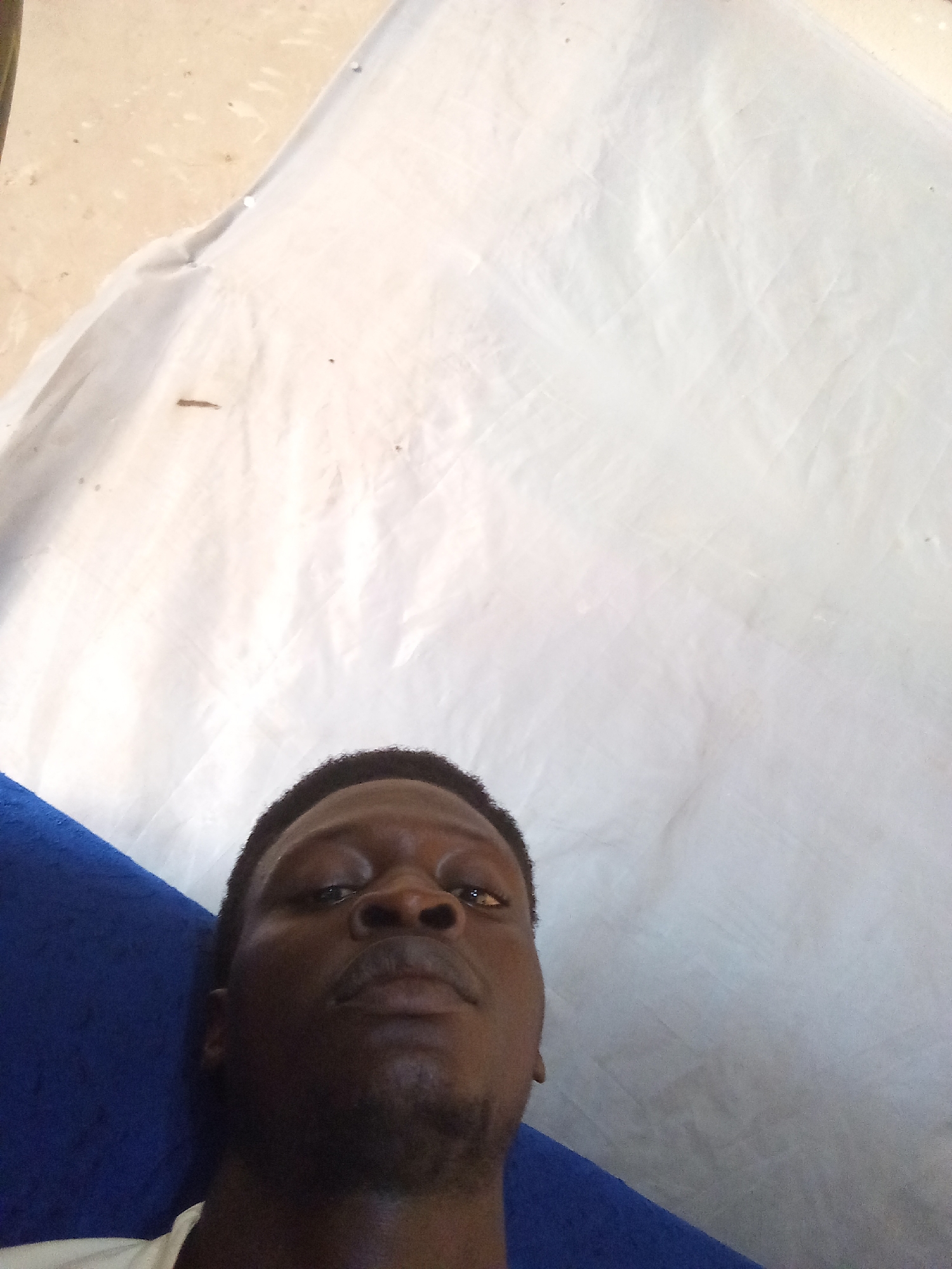 A dark-skinned man is lying down with his head propped up on a blue pillow. He is looking directly at the camera. The background is a white tarp that covers a sandy area. The man's shirt is white and visible at the bottom of the frame. The lighting suggests it is daytime, and the image appears to have been taken indoors or in a shaded outdoor area. The man's facial features are prominent, with dark eyes and a slight stubble on his chin. The overall impression is a casual, possibly candid, photograph.