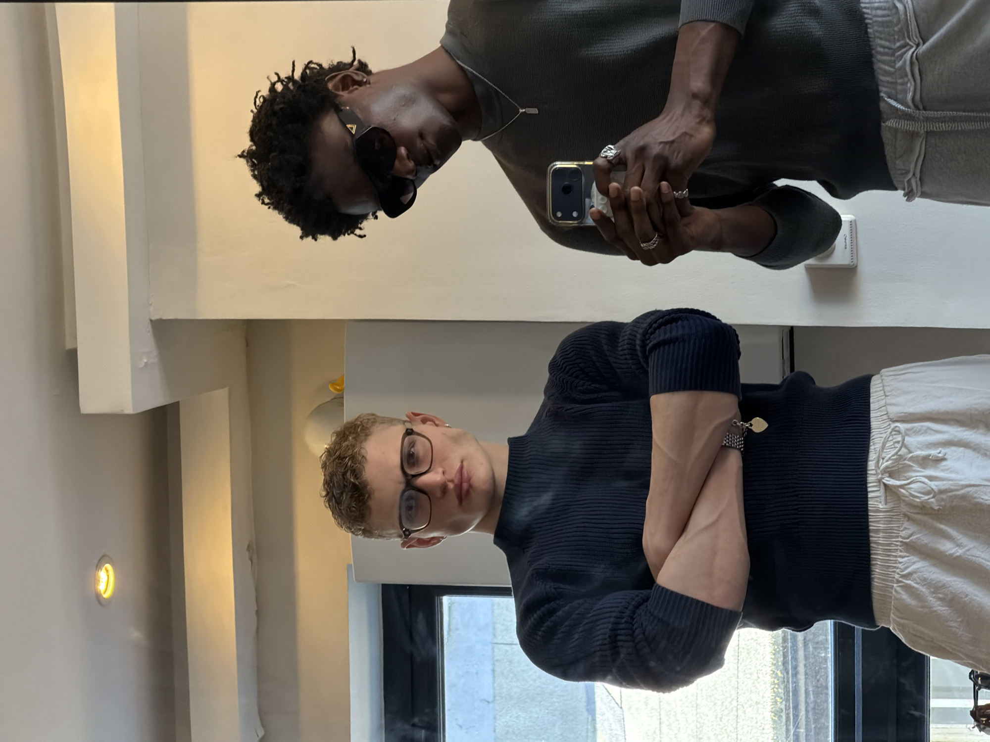 The image shows two young men appearing to take a selfie in a room that may be an apartment or studio in London, United Kingdom. The top man is Black, wearing sunglasses, a grey top, grey sweatpants, and holding a phone in front of his face as if taking a picture. He has rings on his fingers and a necklace. The lower man is White, wearing glasses, a navy blue ribbed sweater, and light-colored pants. His arms are crossed. A window is visible behind him, with an out-of-focus view of a building. The environment shows a modern interior with neutral-colored walls and recessed lighting.
