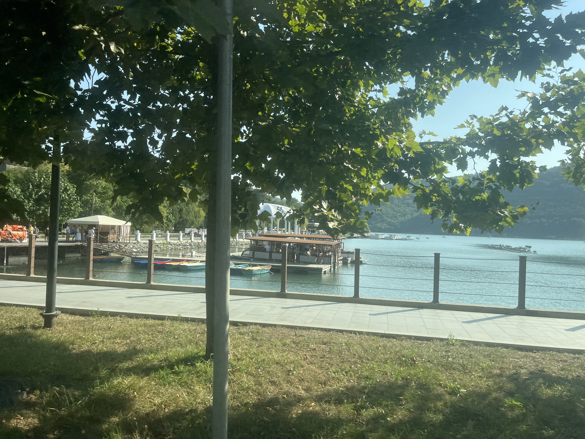 The image shows a waterfront scene under a bright, clear sky, viewed through the branches of lush green trees. In the foreground, a section of a paved walkway runs parallel to a grassy area. A metal railing with horizontal bars separates the walkway from the water. Beyond the railing, the water is calm and reflects the blue sky. Several small boats are docked near a wooden pier, where people are visible. One larger boat, possibly a tour boat, is also moored on the water, with passengers on deck. In the background, hills covered in greenery rise from the far side of the water, and several small white structures are visible along the shoreline. The overall impression is one of a peaceful, recreational area.