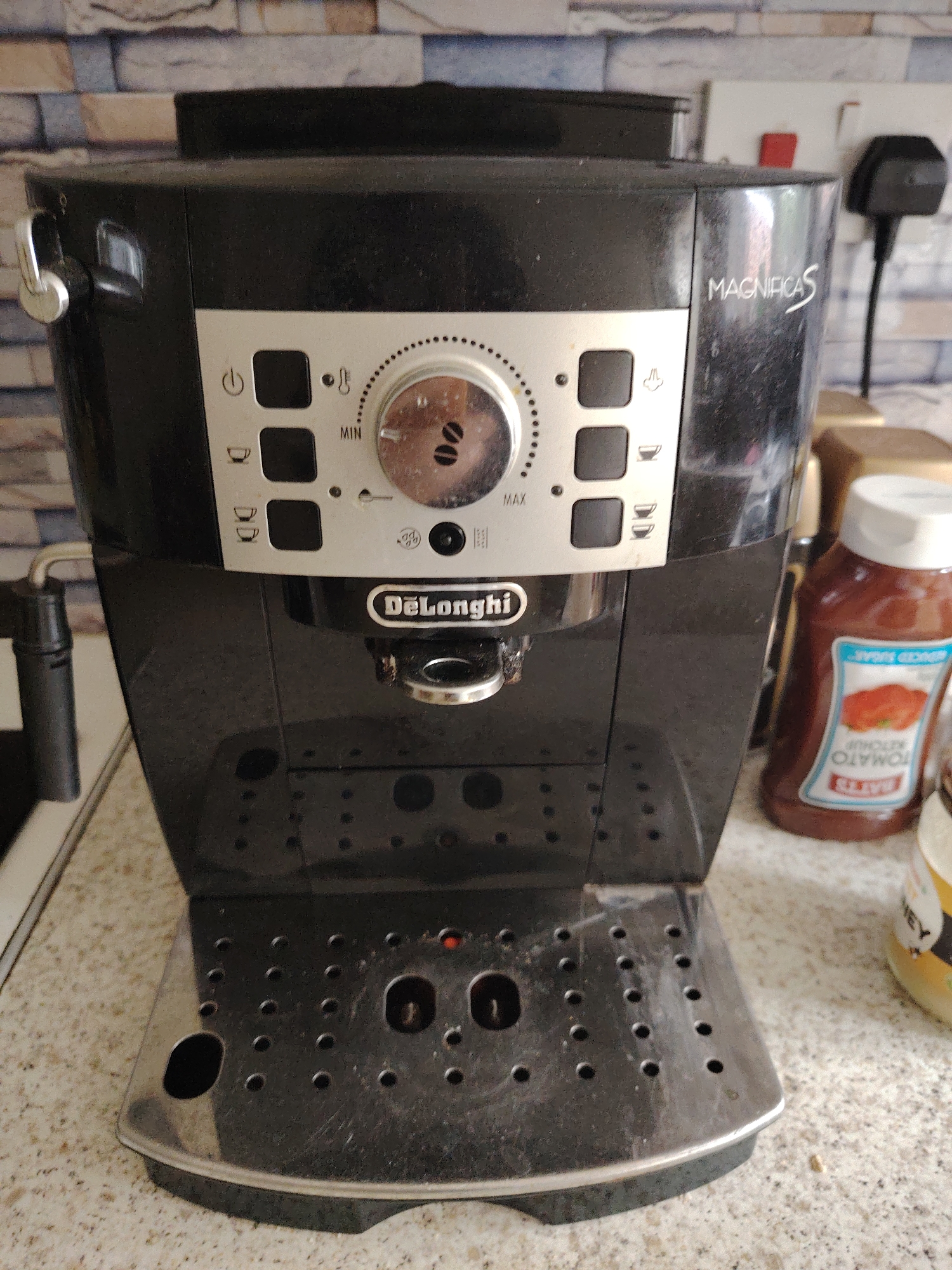Located in Huntingdonshire, United Kingdom, the image displays a black De'Longhi MagnificaS automatic espresso machine situated on a light-colored, speckled kitchen countertop. The machine exhibits signs of use, including dust accumulation on its top surface and sides, and water marks on its perforated metallic drip tray. Its silver control panel features a central rotary dial marked with coffee bean graphics and a scale from 'MIN' to 'MAX'. Surrounding the dial are several square buttons depicting symbols for power, steam/hot water function, single espresso cup, and double espresso cups. The brand name "De'Longhi" is visible above the coffee spouts, and "MAGNIFICAS" is embossed on the machine's right side panel. A chrome steam wand extends from the left side of the unit. The background consists of a tiled wall with a pattern resembling light-toned rectangular bricks in shades of beige, grey, and blue. To the right of the machine, a white electrical wall socket with a red switch is visible, along with a black UK-style power plug. Partially present on the countertop are a brown plastic squeeze bottle identified as "SAINSBURY'S REDUCED SUGAR TOMATO KETCHUP" and a yellow jar with an indistinct label. No individuals are present in the scene.
